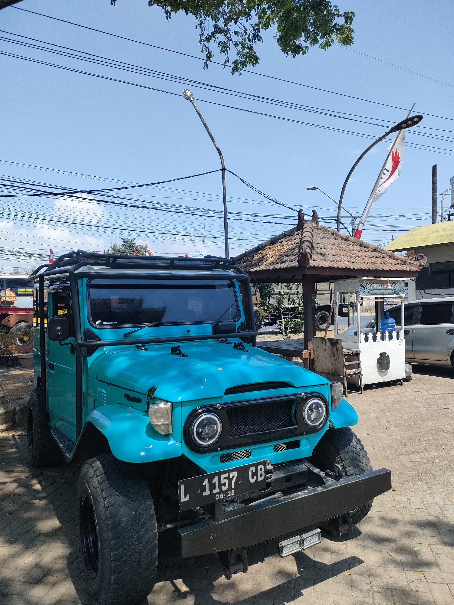 A bright turquoise, off-road vehicle is parked on a paved area outdoors. The vehicle has a black roll cage, a roof rack, and large, knobby tires. The front of the vehicle displays a license plate that reads "L 1157 CB" with "08.26" below it. Behind the vehicle, a small open-air pavilion with a tiled roof stands. Next to the pavilion is a white cart with a blue water jug on top, and a white car is parked to the right of the cart. Power lines and a street lamp are visible in the sky. The scene appears to be located in a public area, possibly a roadside stop or parking lot, in Curahdringu, Indonesia, as indicated by the presence of local flags and the style of the pavilion.