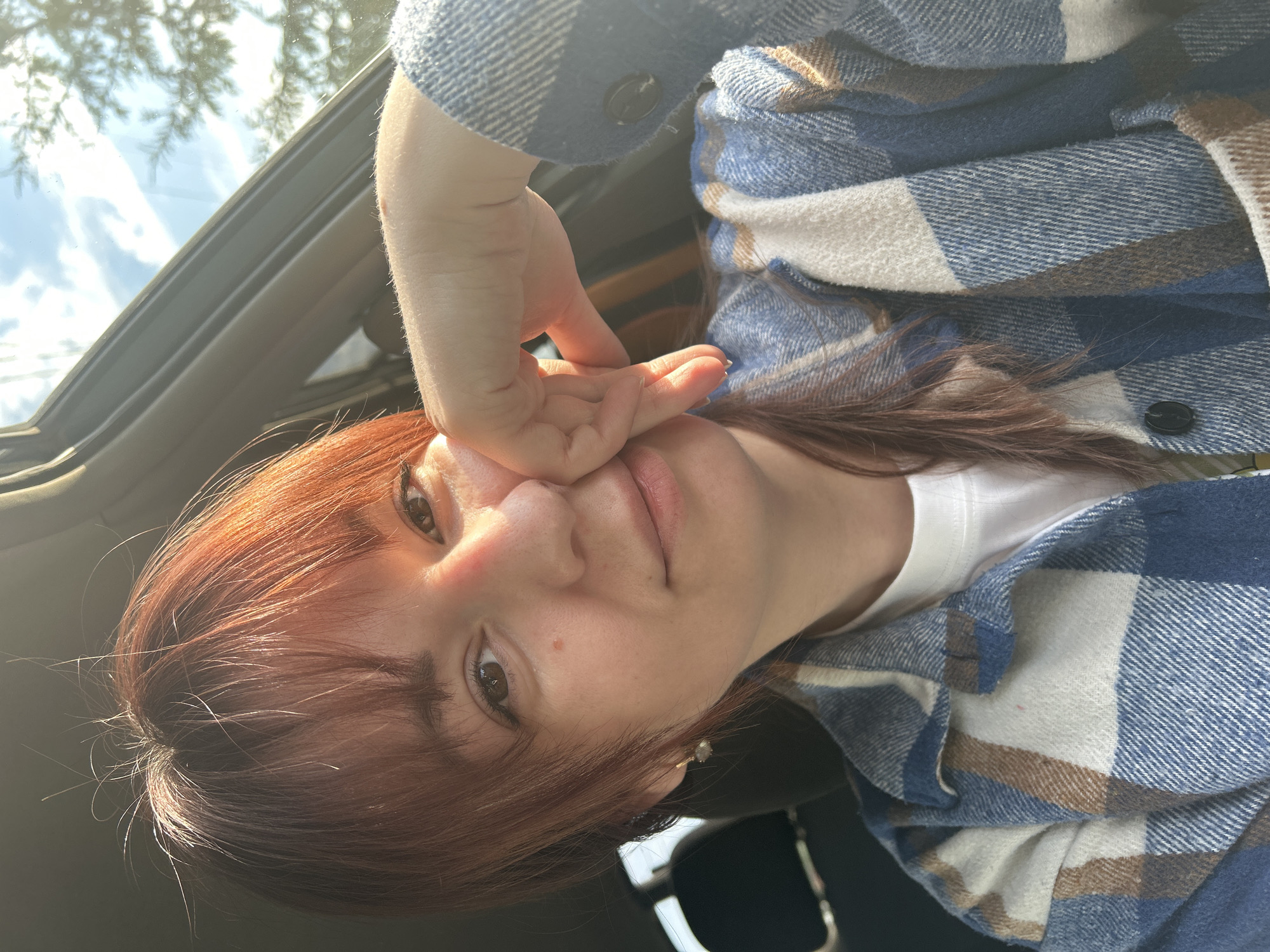 A woman with auburn hair and a blue and white flannel shirt is taking a selfie in a car. Her right hand is near her face. The car's sunroof is open, showing sky and trees. The location is not possible to determine from the image.