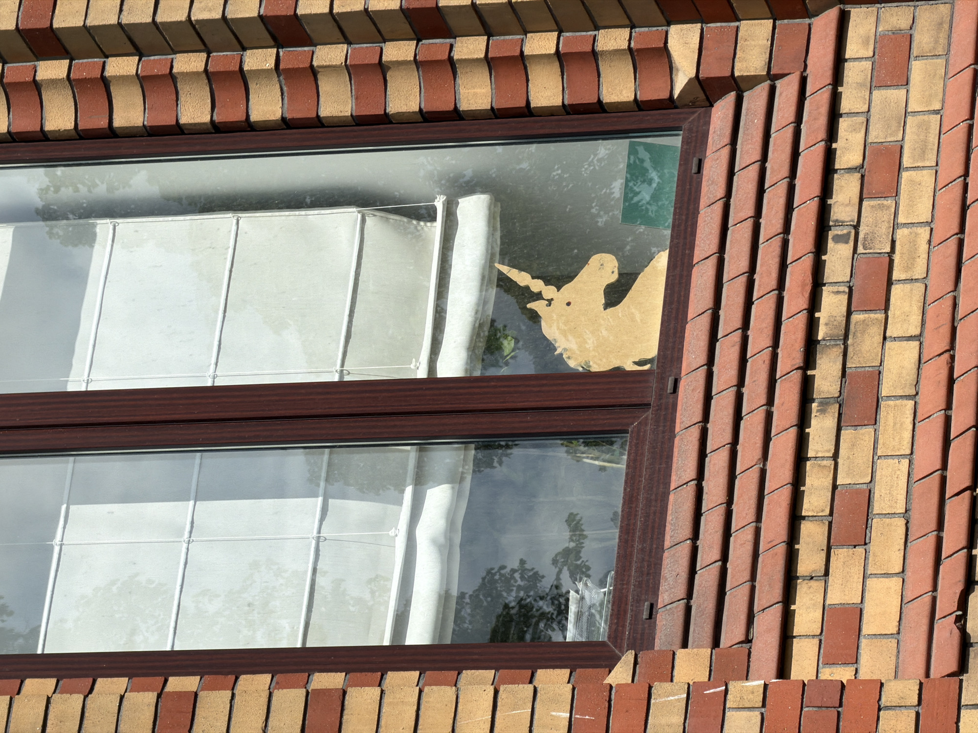 A close-up shot captures a section of a building's exterior, centered on a rectangular window with a dark brown frame. The building's facade is constructed from bricks, laid in alternating patterns of reddish-brown and light beige/yellow. Above and below the window, the bricks are arranged horizontally, creating a wavy, textured effect. To the right of the window, the bricks are predominantly laid vertically, forming distinct, angled rows.
Inside the window, light-colored, possibly white or cream-colored, vertical blinds or curtains are partially drawn across both the upper and lower panes. Reflected in the glass, the faint outlines of trees or foliage are visible, suggesting an outdoor setting on a sunny day. A striking detail is a cutout of a mythical creature, likely a unicorn with wings, affixed to the inside of the top-right window pane. This cutout, in a light yellow or golden hue, faces left, with its horn and part of its wings clearly visible. A faint green rectangular shape is also visible in the upper right corner of the same pane.
The scene is brightly lit by natural sunlight, casting strong shadows that highlight the relief and texture of the brickwork. No people or visible text are present. The overall impression is one of an urban architectural detail, with the unicorn cutout adding a whimsical, personal touch to the otherwise functional window. This building is located in Saint Petersburg, Russia.