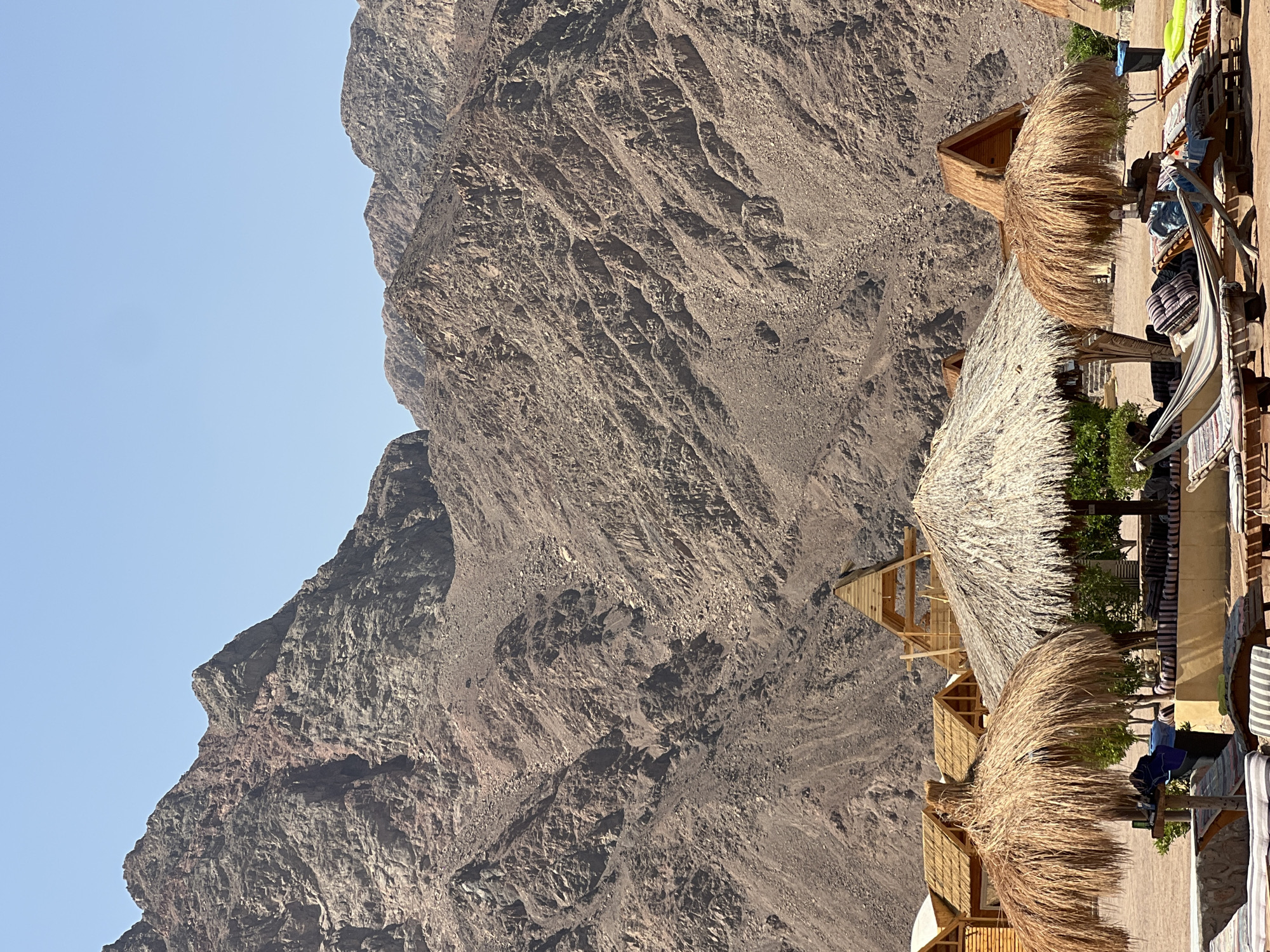 This is a medium shot, eye-level, outdoor photograph taken on a sunny day. The scene depicts a resort area with several thatched-roof structures and a hammock in the foreground, set against a backdrop of rugged, rocky mountains. The mountains are large and imposing, with varying shades of brown and gray, and appear to be a prominent geological feature of the area.

In the foreground, there are lounge chairs with striped cushions, a hammock strung between two wooden posts, and two large thatched umbrellas providing shade. The umbrellas are made of dried palm fronds. Behind these, several small wooden cabins with thatched roofs are visible. One of these cabins, in the center of the image, has an A-frame structure made of unfinished wood, suggesting ongoing construction or a specific architectural style. The ground appears to be sandy.

The lighting suggests it is daytime, with harsh shadows indicating a bright sun. The sky is clear and a pale blue. There is no visible water, although the presence of beach-like amenities suggests proximity to a body of water, likely the Red Sea, given the location.

The image conveys a sense of a tropical or desert resort environment. The architecture, with its use of natural materials like thatch and wood, is characteristic of beach or eco-tourism accommodations. The overall impression is one of a relaxed, vacation-oriented setting in a remote, natural landscape. The location is the city of Nuweiba, Egypt, a popular tourist destination known for its beautiful beaches and proximity to the Sinai Mountains.