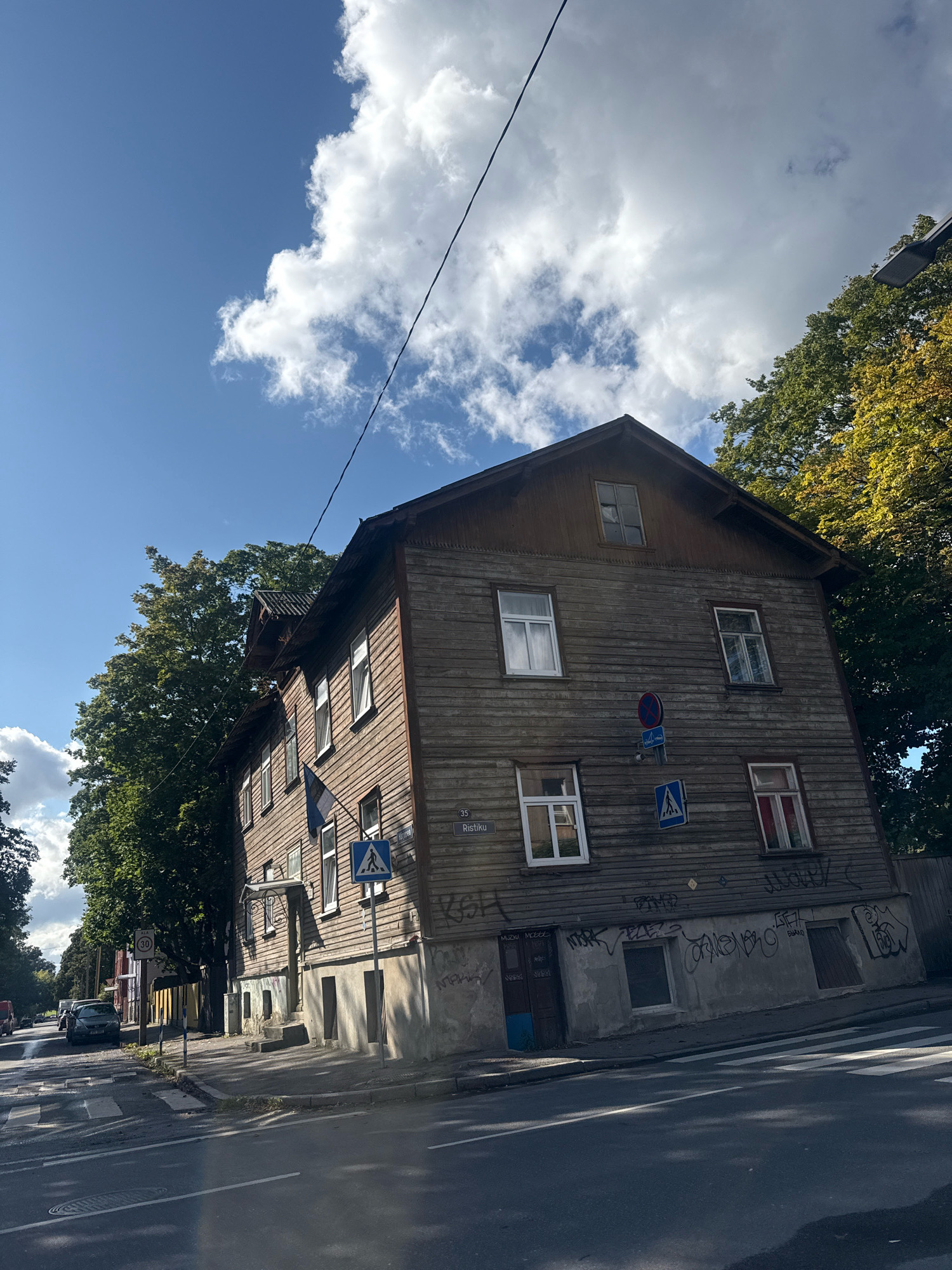 The image shows a wooden residential building on a street in Tallinn, Estonia, with a blue sky and white clouds overhead. The building has multiple floors, visible windows, and a gabled roof. There are trees on either side of the building. A street with a crosswalk is in the foreground. Traffic signs are visible on the building and on the street. The building appears to be in an urban or suburban area. There are no people visible in the image. The overall scene depicts a day in Tallinn with a focus on the architecture and streetscape.
