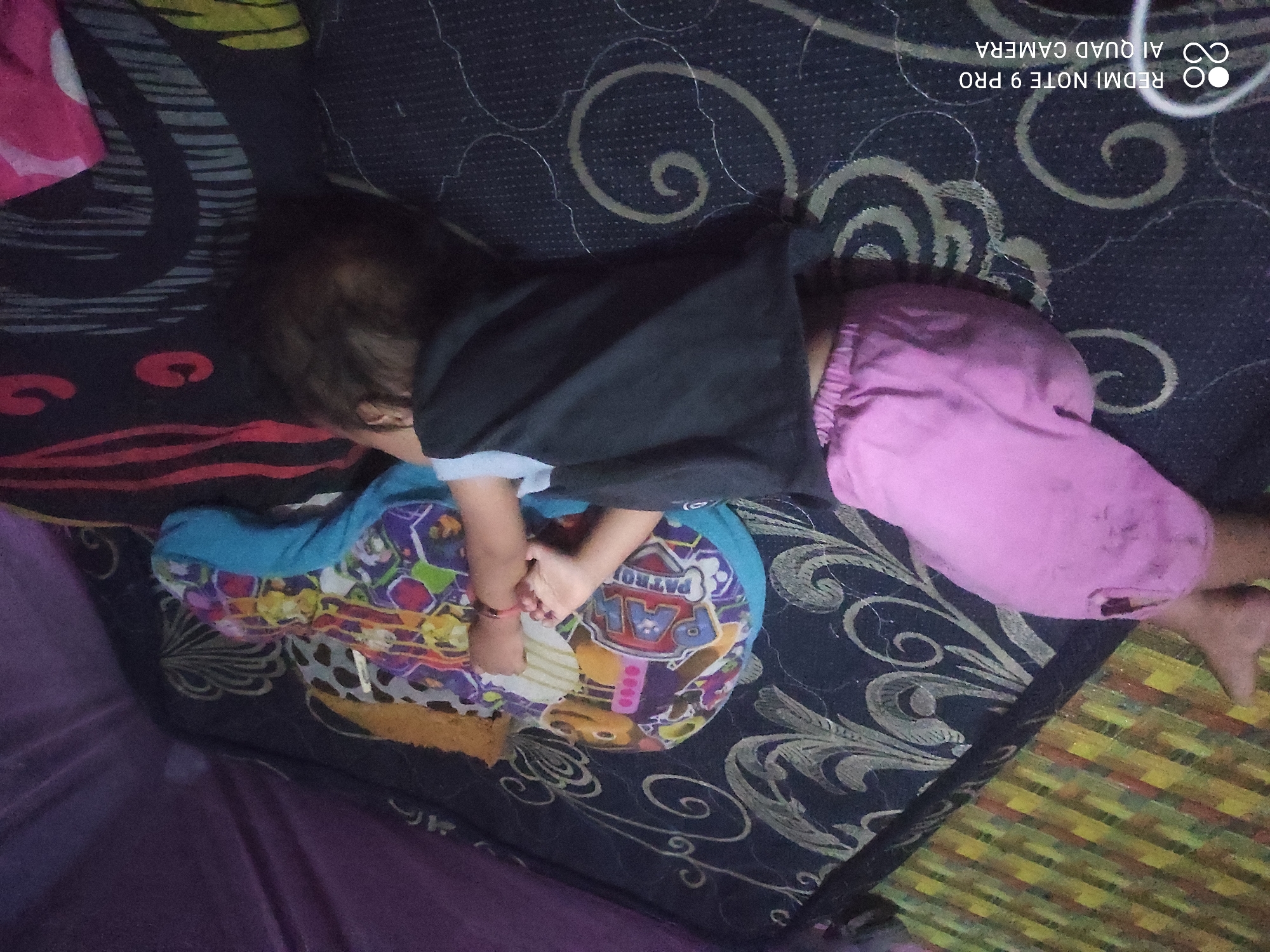 A child sleeps on a mattress with a dark, patterned fabric. The child wears pink shorts and a dark t-shirt. A Paw Patrol pillow is beside the child. The room is dimly lit. The watermark "REDMI NOTE 9 PRO AI QUAD CAMERA" is visible in the lower left corner. The image does not contain information about the location in the city of Sakra, Indonesia.