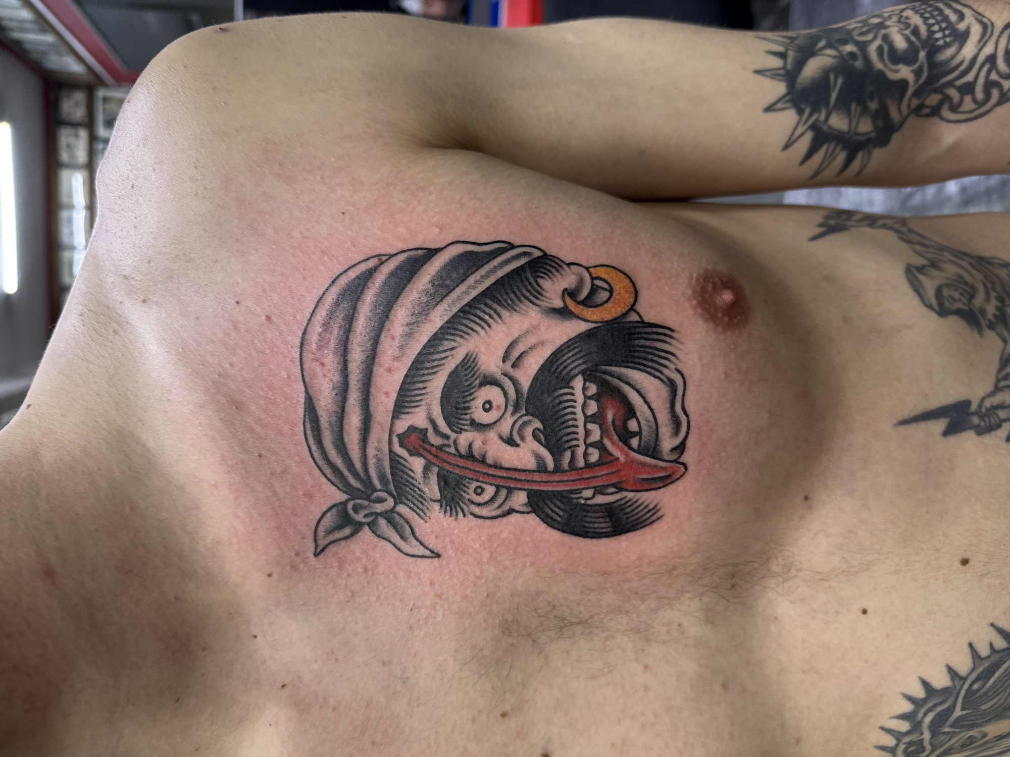 A torso with pale skin is in the center of the image, featuring a tattoo on the upper chest. The tattoo depicts a monstrous face with a red tongue sticking out, and a bandana tied around its head. An orange ring is pierced through what appears to be a nostril. There are other tattoos visible on the person's arms and upper chest, including a creature with sharp spikes and teeth on the arm. The background is somewhat blurred, but appears to be an indoor space with lighting fixtures on the ceiling. The image has no indication about its origin in Moscow, Russia.