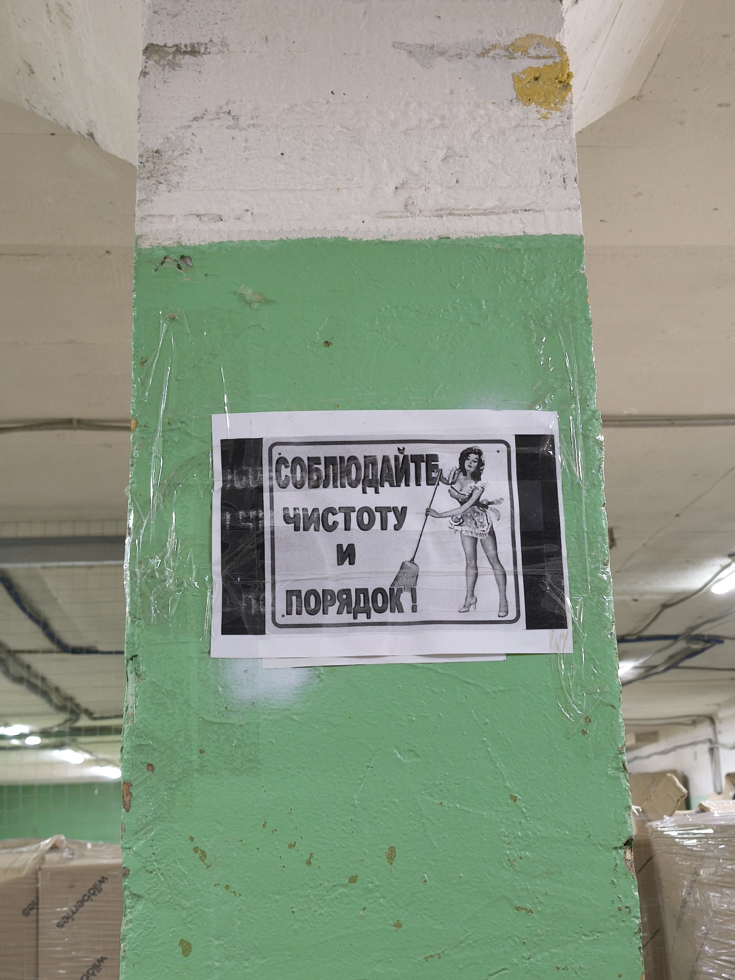 A black and white sign is adhered to a green painted concrete pillar with clear tape. The sign has a black border and text in Russian, "СОБЛЮДАЙТЕ ЧИСТОТУ И ПОРЯДОК!" which translates to "MAINTAIN CLEANLINESS AND ORDER!". A pin-up style illustration of a woman in a short dress and high heels holding a broom is on the right side of the sign. The pillar is painted white above the green. The location is identified as Moscow, Russia based on the Russian writing. The pillar appears to be inside a warehouse with visible boxes stacked and covered in plastic wrap in the background. Exposed wiring and ceiling lights are also visible in the background.