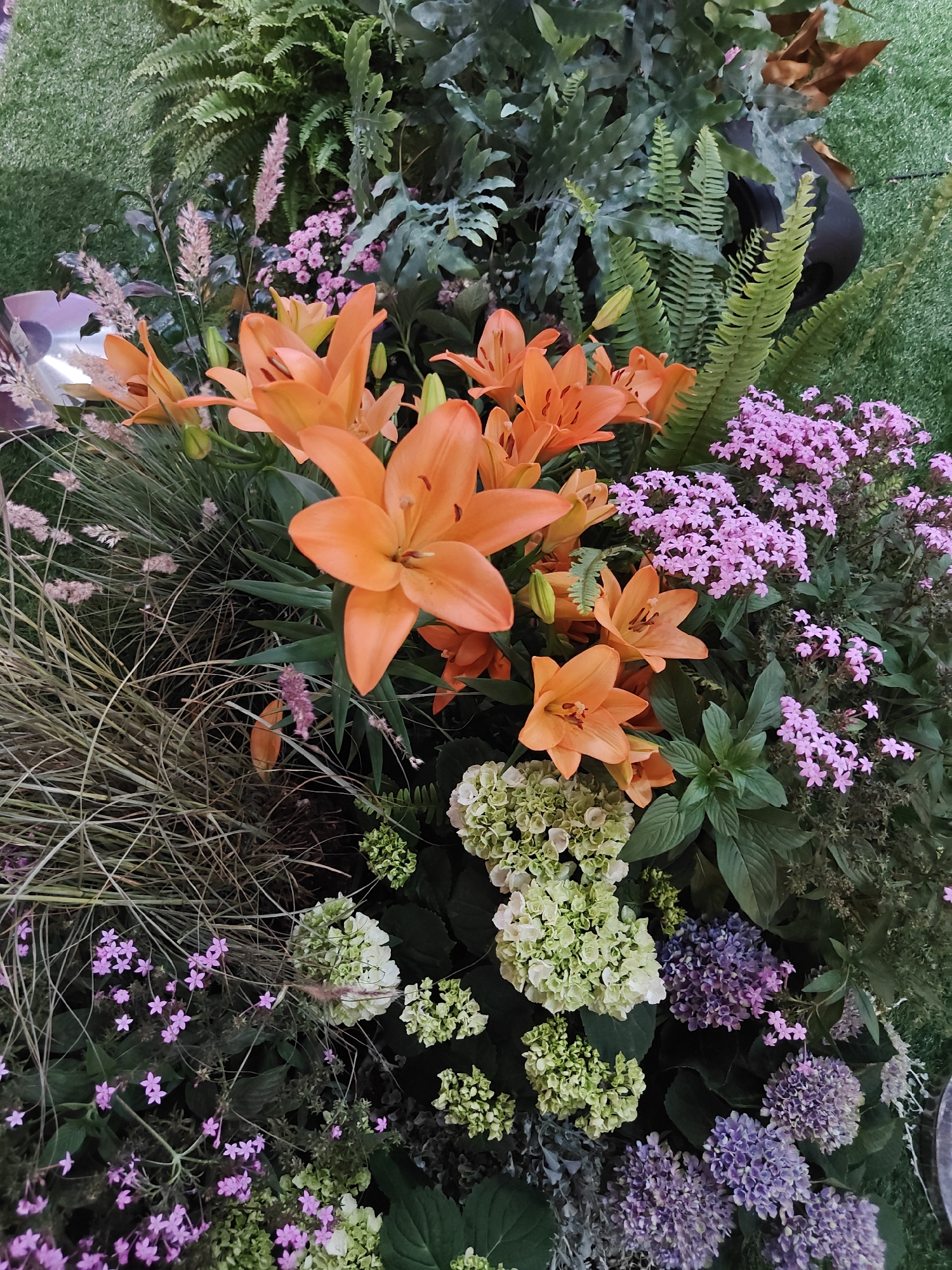 The image is a close-up, high-angle shot of a vibrant floral arrangement. Dominating the foreground are several large, bright orange lilies with prominent stamens and dark centers. Interspersed with the lilies are clusters of small, delicate pink flowers, and feathery, pale pink plumes. To the left and center, a mass of thin, straw-colored dried grass provides textural contrast. In the lower portion of the image, there are groupings of pale green and purple hydrangeas, their florets tightly packed. Behind the flowers, various types of green foliage are visible, including broad leaves and delicate fern fronds, all set against a backdrop of what appears to be artificial green turf. The lighting is even and diffused, highlighting the rich colors and intricate details of the plant life. No people or actions are depicted. The context of Tel-Aviv, Israel, is not visually present in the image itself.