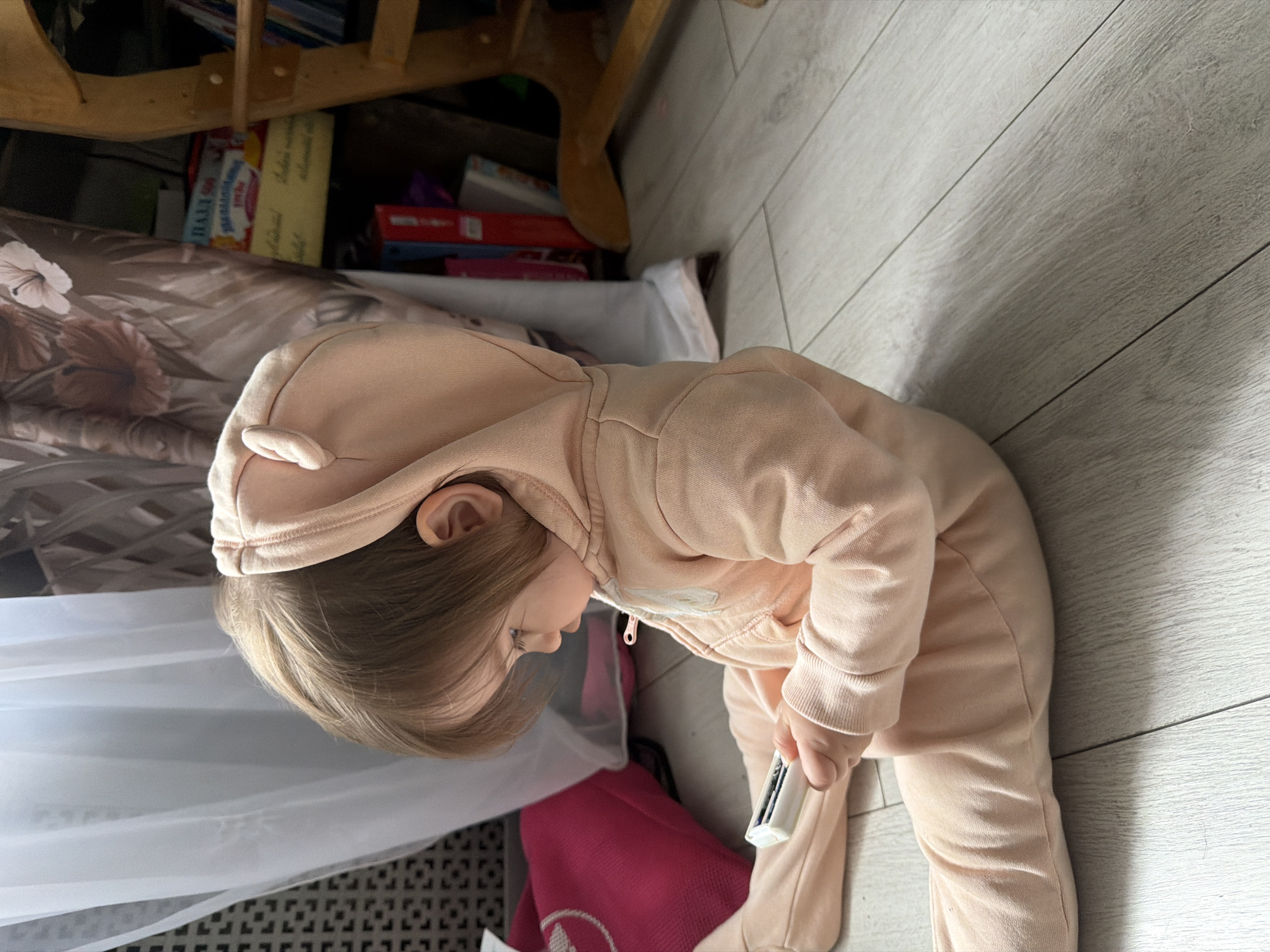 A young child wearing a pink hooded jumpsuit stands on a light gray wooden floor holding a white rectangular object with a black element. The child is bending over and appears to be looking at something near the floor. Behind the child are portions of furniture and items, including a light-colored wooden frame with a shelf, a patterned surface resembling bedding, and a pink object. The location cannot be confirmed as Novosergievka, Russia, based on the image content.