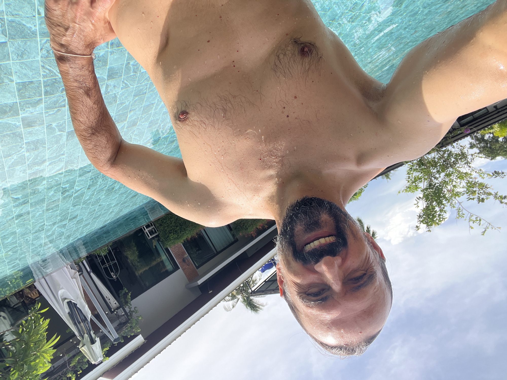 A selfie taken in a swimming pool in บ้านบางรักษ์, Thailand, shows a man with a beard and receding hairline smiling at the camera. The pool's blue tiled surface is visible in the upper left of the frame. In the background, foliage and a building with a white roof are visible. The sky is partly cloudy. The man is shirtless.