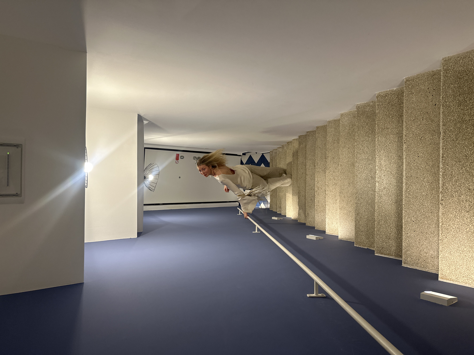 The image shows a person in a white outfit suspended horizontally above a blue floor, seemingly defying gravity. The scene is set in an interior space with walls and a ceiling, suggesting a room that has been rotated to create an optical illusion. To the right of the person, there are vertical structures that resemble stairs. A white railing runs horizontally along the bottom of these structures. The illumination is provided by multiple sources, including a bright light in the distance and some light sources near the floor. Behind the person, there is a wall with drawings of fan-like structures, and an exit sign. The image is taken at the Museo Archeologico Luigi Bernabò Brea in Lipari, Italy.