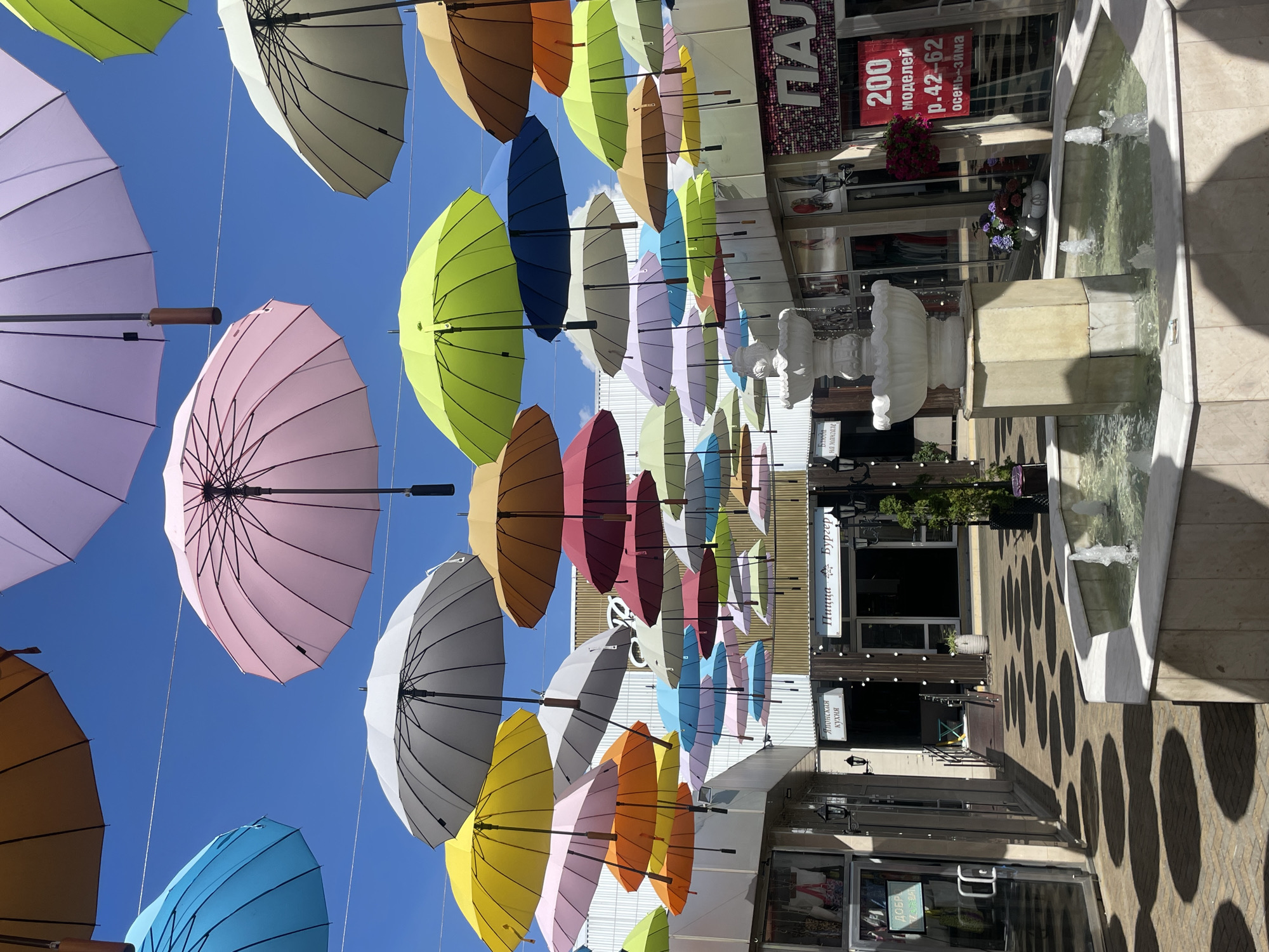 A street view shows an outdoor pedestrian walkway lined with shops and decorated with a multitude of colorful umbrellas suspended overhead. A multi-tiered fountain is centrally located in the walkway, with water spouting from its various levels. The shops appear to be in Sochi, Russia, with signage in Russian visible on some of the establishments. The umbrellas cast circular shadows on the paved ground, creating a patterned effect. The sky is clear and blue, suggesting fair weather. There are no people visible in the scene.