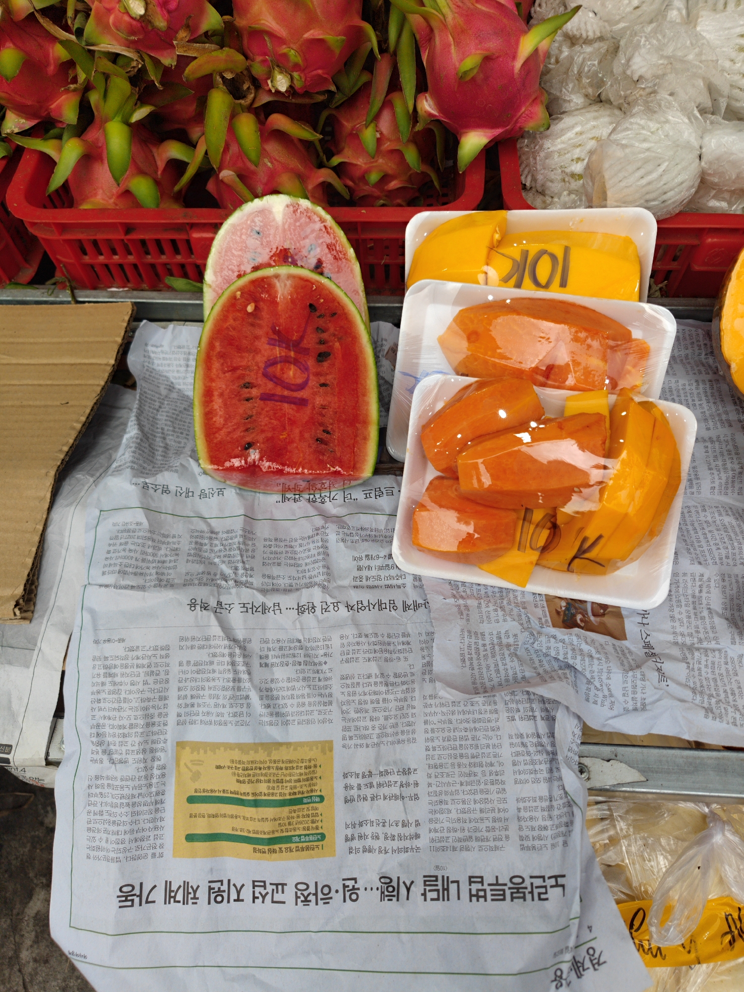 This is a close-up shot of a fruit stand in Cam Ly - Đà Lạt, Vietnam. Dragon fruit is visible in a red plastic basket. Slices of watermelon, wrapped trays of orange papaya, and packaged pomelos are arranged on top of a Vietnamese newspaper spread out on a table. The fruit is priced with the number "10K" written on them, presumably meaning 10,000 Vietnamese Dong. The focus is on the presentation and sale of pre-cut, portioned fruit.