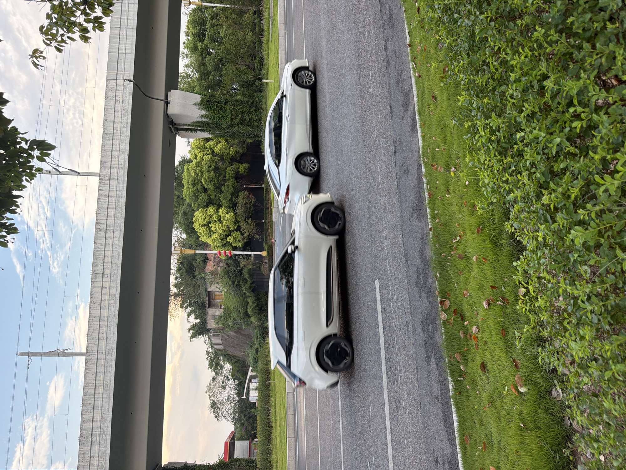 Two white cars are traveling on a multilane road in Jiangjin, China. The car in the foreground is a white SUV, and the car in the background is a white sedan. Both cars are moving in the same direction. The road is paved and has white lane markings. On the left side of the road, there is a grassy area with some fallen leaves. On the right side of the road, there is more grass and some bushes. In the background, there is an elevated structure, likely an overpass or bridge, with train tracks on top. Trees and buildings are visible beyond the overpass. The sky is partly cloudy.