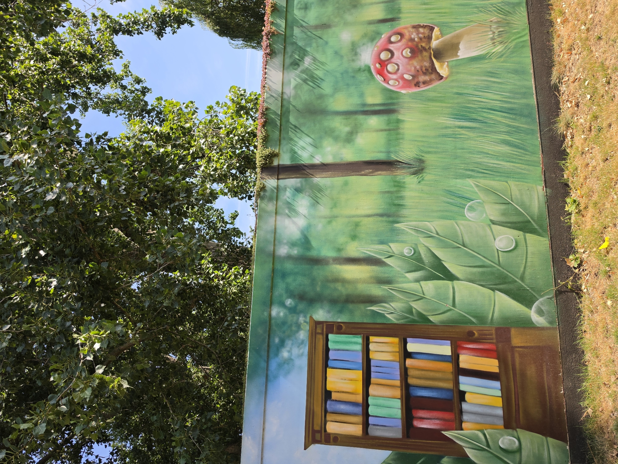 The image shows a mural painted on a wall, depicting a fantasy forest scene. On the left, a bookshelf filled with colorful books is painted. To the right of the bookshelf, large green leaves with dew drops are shown. Further to the right, a red mushroom with white spots is painted in a grassy field. Trees and a blue sky are depicted in the background. The mural appears to be located outdoors, with grass and dirt in the foreground. This mural is located at the Rijswijkse Schouwburg, a theatre in the city of Rijswijk, Netherlands.
