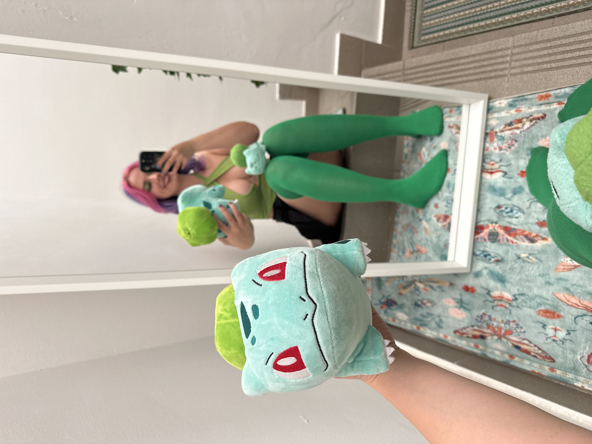 A vertically oriented photograph depicts a woman in green stockings taking a selfie in a mirror while holding a Bulbasaur plush toy. The reflection in the mirror shows her face, phone, a green top, black shorts, green stockings, and another Bulbasaur plush toy. The setting is indoors, evidenced by the walls and flooring. A rug with a butterfly and insect pattern is partially visible near the mirror. There is a flight of stairs adjacent to the mirror. Although I can describe the elements of the image, I cannot determine the location in City of Zagreb, Croatia.