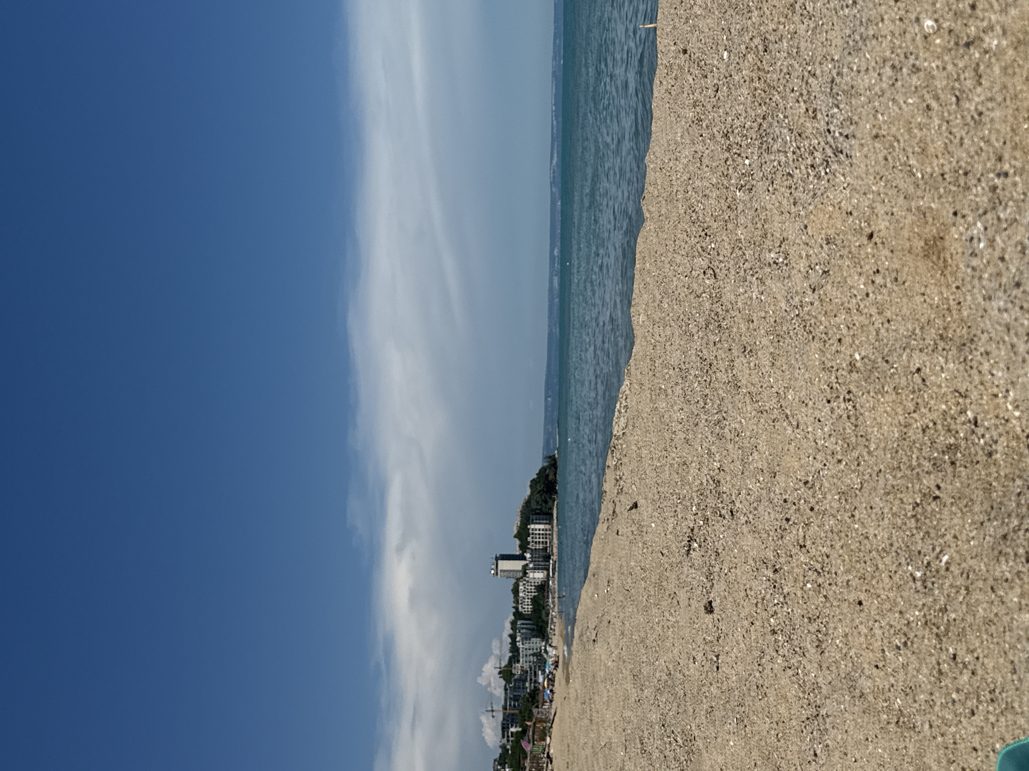 A wide shot of a sandy beach in Varna, Bulgaria, with the Black Sea stretching out to the horizon. The sand in the foreground is coarse and light brown, with scattered pebbles and debris. In the mid-ground, there is a shoreline with several beach umbrellas and a few people enjoying the day. Beyond the beach, a strip of buildings, including apartment complexes and a tall hotel, are visible along a green, hilly coastline. A construction crane can be seen among the buildings on the left. The sky is a clear blue with a few wispy white clouds. The water is a deep turquoise, with gentle waves lapping at the shore. The scene appears calm and sunny.