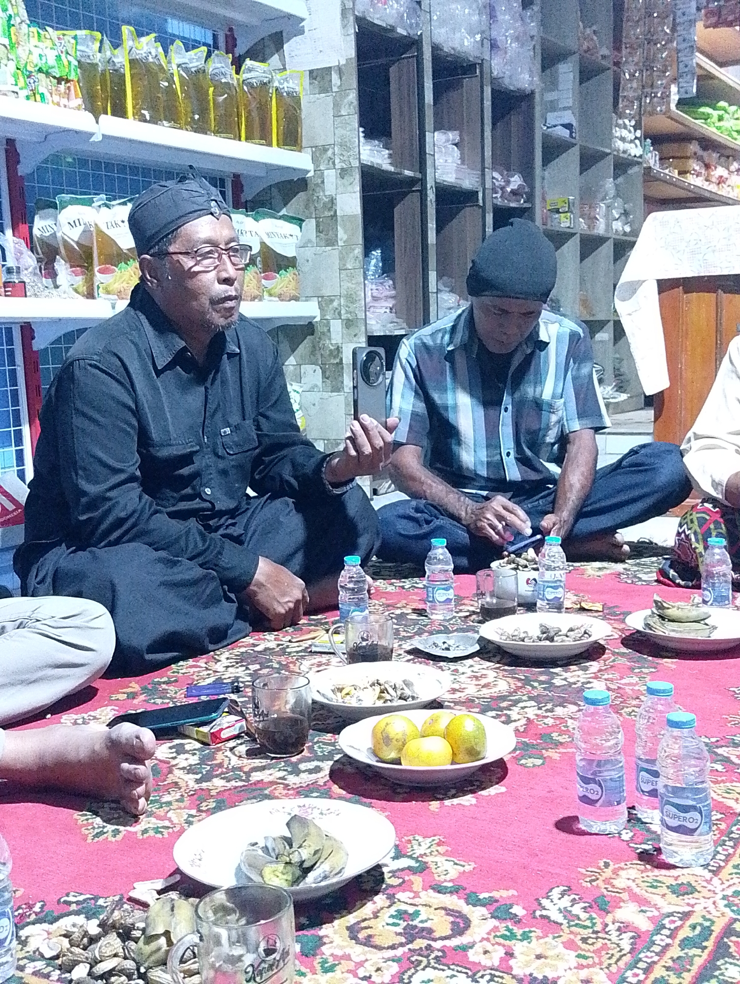 The image depicts an indoor scene, likely in a domestic setting in Cilacap, Indonesia, with shelves filled with packaged goods visible in the background. In the foreground, several individuals are seated on a red carpet patterned with floral motifs. Two men are the most prominent. The man on the left wears a black, long-sleeved shirt and pants, a black cap, and glasses. He is holding a smartphone in his right hand and appears to be looking at it. The man on the right wears a blue and white checkered shirt and jeans. He is looking down at a smartphone he is holding and appears to be interacting with it. Several plates of food, some containing what appear to be nuts and others fruit, are arranged on the carpet, along with glasses of dark liquid and bottles of water. The overall atmosphere suggests a casual gathering or meal.
