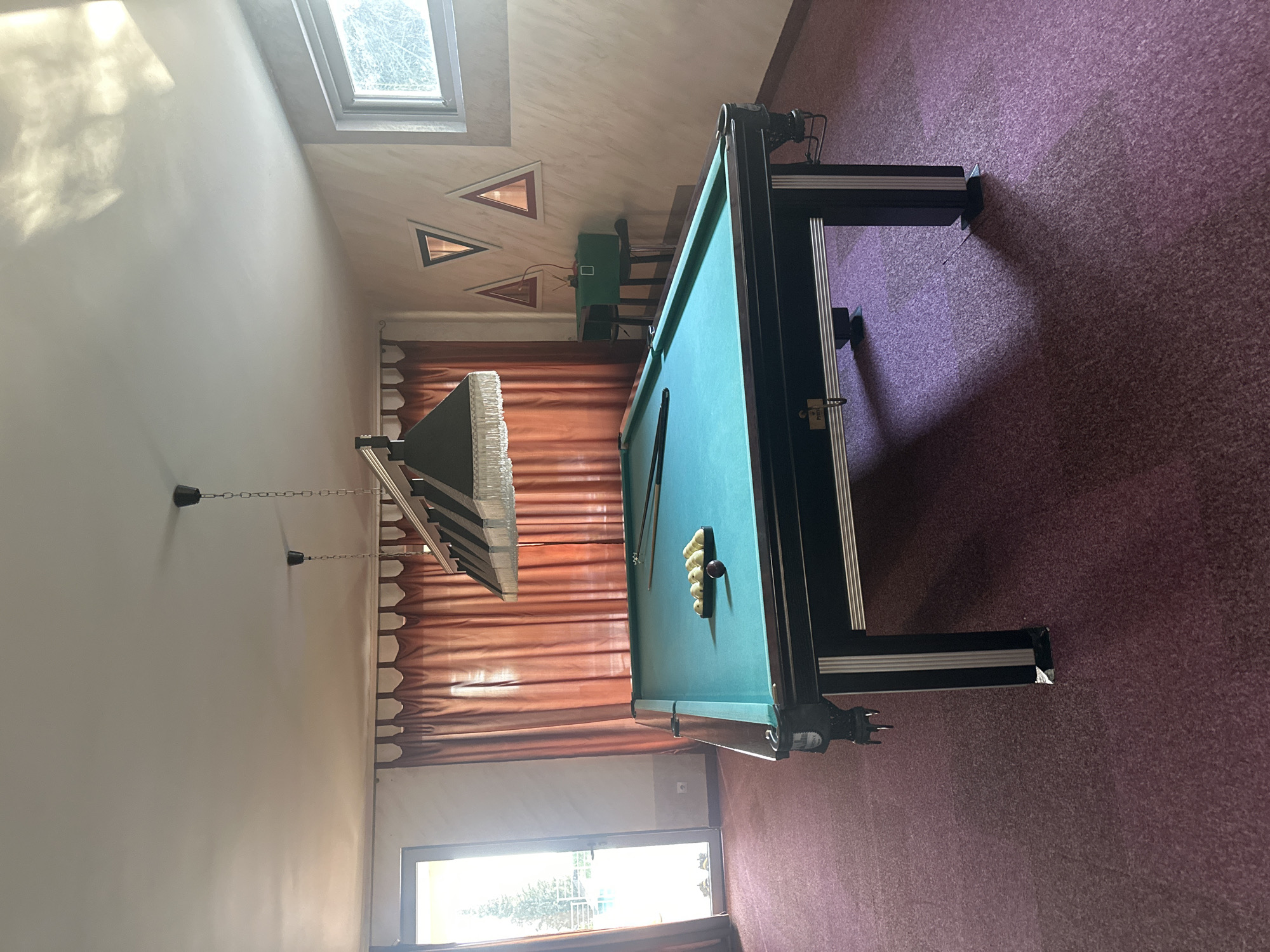 The image depicts a room in a private residence or establishment, likely a game room, located in Brca, Montenegro. The central object is a green pool table with black trim and legs, positioned on a purple carpeted floor. Several billiard balls, including a red one and a rack of white balls, are placed on the table. Above the pool table, a multi-lamp lighting fixture hangs from the ceiling. In the background, there are large, rust-colored curtains covering a window and a glass door that leads outside to a bright area with trees and a railing. The walls are painted in a light, textured color, with several triangular decorative frames on one wall. To the right, a small green cabinet sits on a stand near a window with a view of trees. The room is illuminated by natural light entering from the door and window, as well as the overhead lamps.