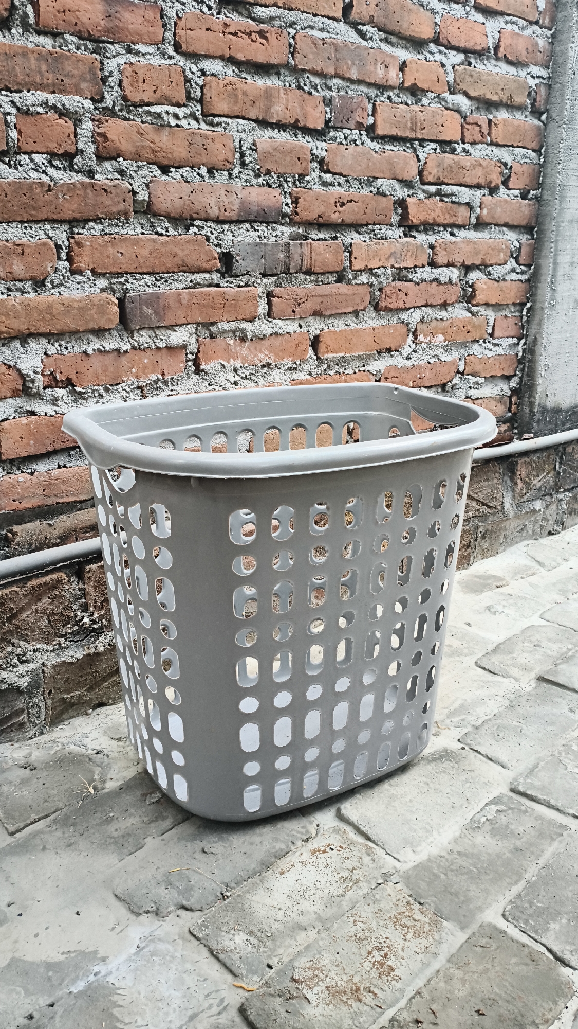 In Kerep Kidul, Indonesia, a grey plastic laundry basket stands on a paved area in front of a brick wall. The basket is positioned in the center of the frame, facing slightly to the left. It is empty and has a perforated design with many oval-shaped holes. The brick wall behind the basket is made of reddish-brown bricks with visible mortar. To the right of the brick wall, a section of a concrete structure is visible. The ground is covered with rectangular paving stones that appear weathered and have some dirt and debris on them. The lighting suggests it is daytime, casting subtle shadows on the ground. No people are present in the image.