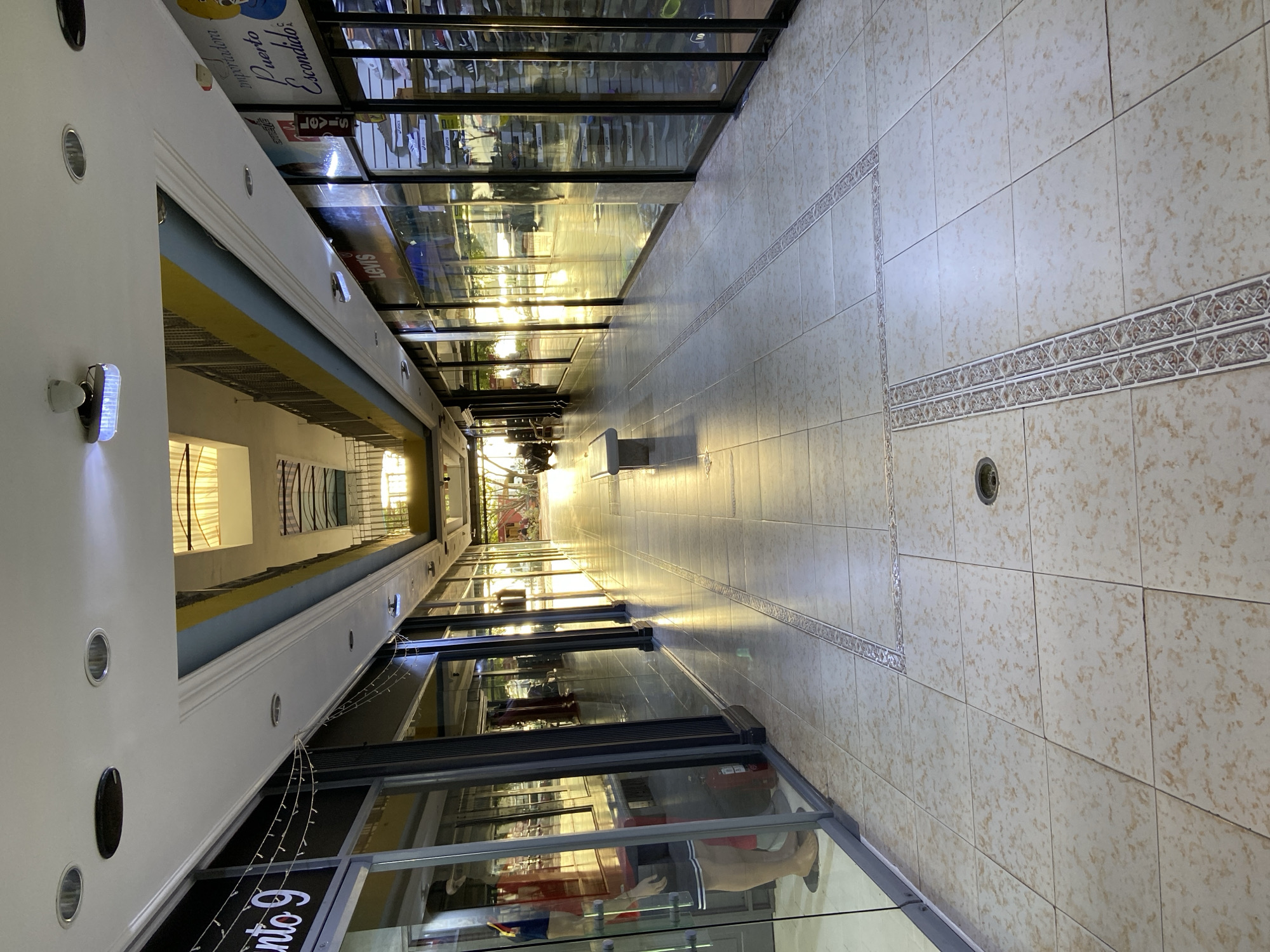 The image shows a well-lit, interior hallway of a commercial establishment, likely a shopping arcade or mall, in Juan Griego, Venezuela. The hallway is lined with storefronts featuring large glass windows, revealing merchandise and mannequins. The floor is tiled with a light-colored ceramic tile with a decorative border pattern running through the center of the hallway. Natural light streams in from the far end of the hallway, creating a warm, sunlit atmosphere. On the left side of the hallway, a store window displays a mannequin dressed in athletic wear, with a red fire extinguisher visible inside the store. A bench is positioned in the middle of the hallway on the left side, providing a place to sit. The ceiling is modern, with recessed lighting and some decorative architectural elements. The overall impression is one of a clean, organized, and inviting commercial space.