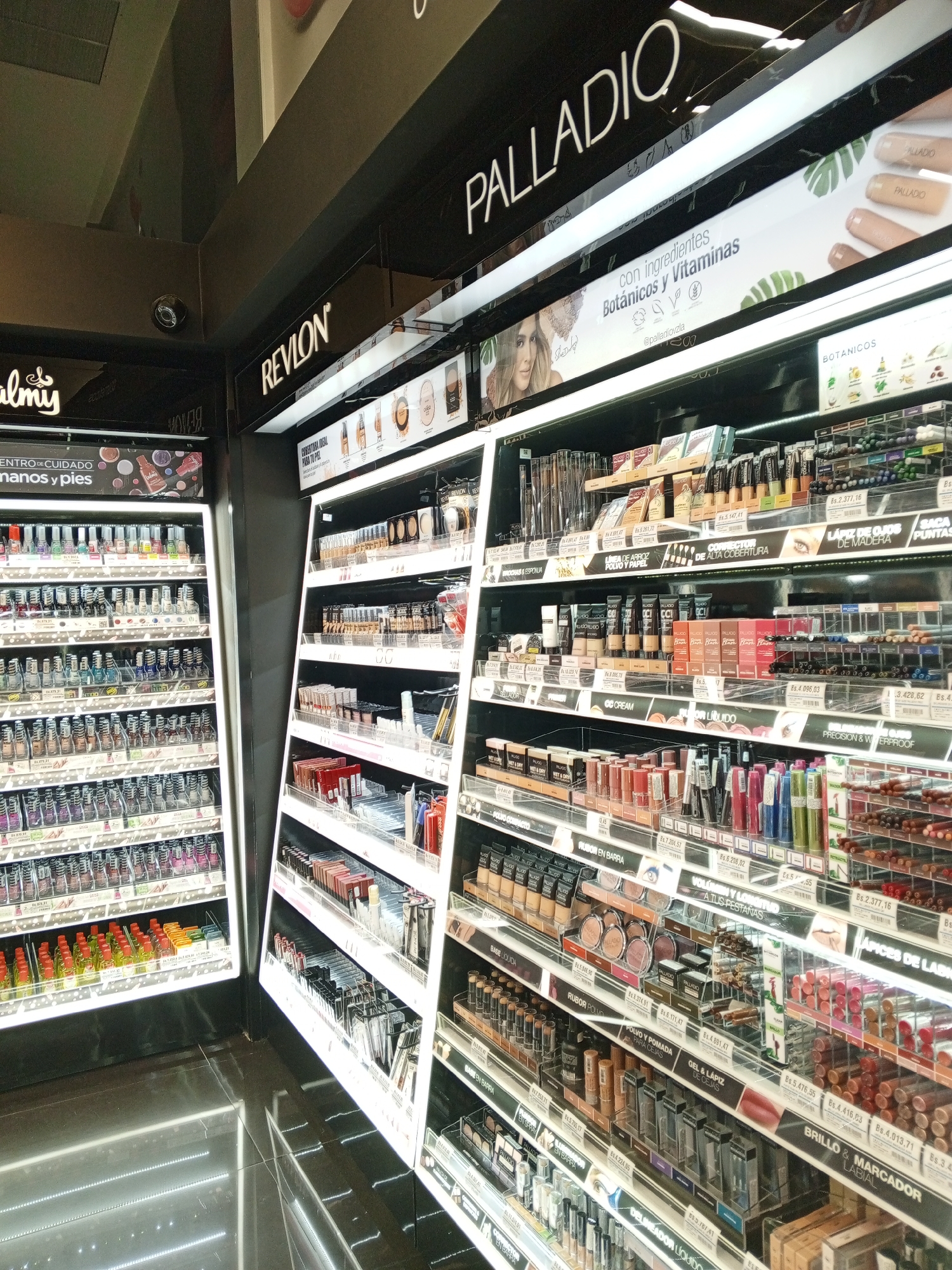 The image depicts a well-lit interior shot of a retail store, specifically a beauty and cosmetics section. Two main shelving units are visible. One is stocked with nail polishes and other hand/foot care products, with the brand name "Palmy" displayed at the top. The other unit is divided into sections for brands like Revlon and Palladio, displaying a range of cosmetic products including foundation, eyeshadow, lipsticks, and eyebrow pencils. Each section has a price display in Bolivars (Bs.) written on it. The scene is characterized by organized shelving, a variety of cosmetic products, and illuminated product displays. Given the context of the image, the location of the store is likely in Pampatar, Venezuela.