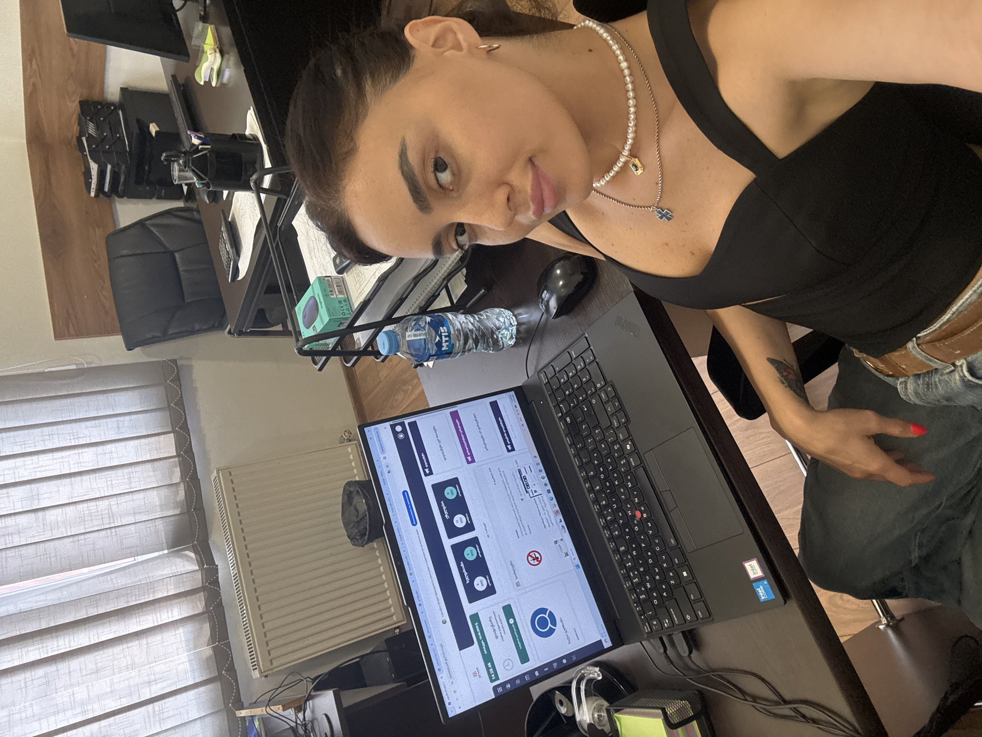 A young woman with dark hair, wearing a black top and jeans, is seated at a desk in what appears to be a home office. She is looking towards the camera with a slight smile. On the desk, there is a laptop displaying a web page with Georgian text and icons, a water bottle, and a computer mouse. In the background, there are blinds, a radiator, and office equipment including monitors and chairs. The scene suggests a work-from-home or study environment. The lighting indicates it could be daytime. The visible text on the laptop screen includes Georgian characters and what looks like a timer displaying "14:28:46". The woman is wearing a pearl necklace with a pendant.