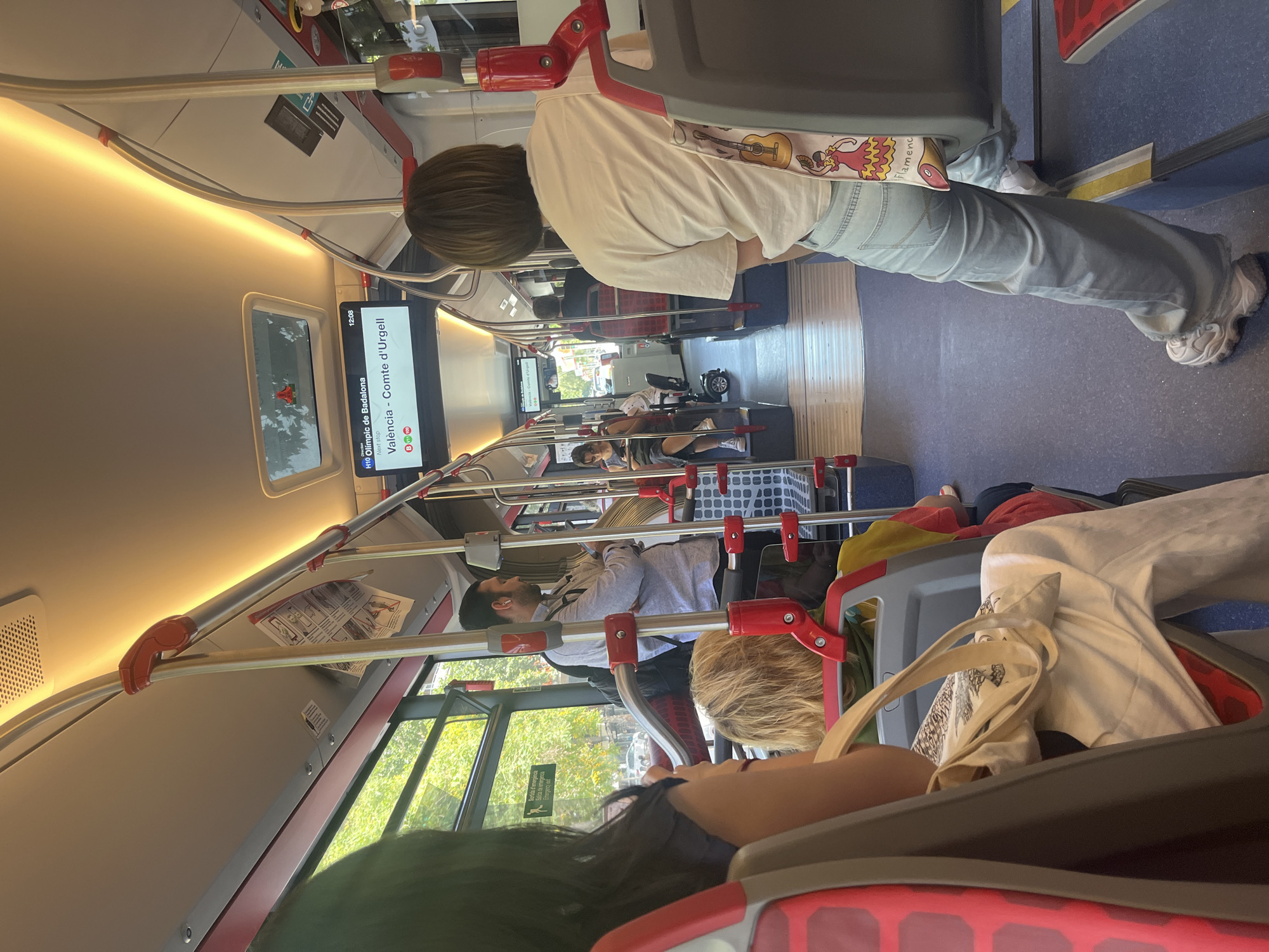 The image shows the interior of a bus in Barcelona, Spain, during the daytime. The bus appears to be in motion, with passengers seated and standing. A digital display screen at the front of the bus shows the current destination as "Valencia - Comte d'Urgell" and the time as "12:08". The interior of the bus is well-lit with overhead fluorescent lighting. There are red and gray seats, metal poles for passengers to hold onto, and large windows offering a view of green trees and city streets outside. Several passengers are visible, including a woman in the foreground with long dark hair, a woman with blonde hair, a man in a light blue shirt standing and looking at his phone, and other passengers further back. The overall atmosphere is that of a typical public transportation scene in a city.