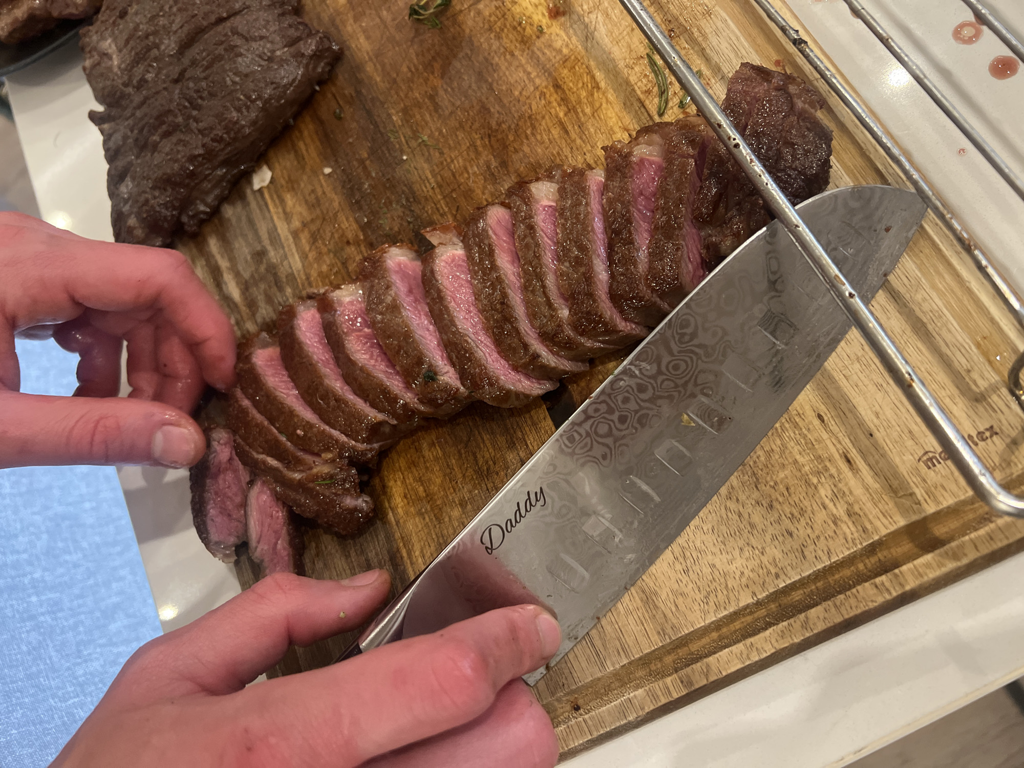 The image displays a close-up view of a person carving a cooked steak on a wooden cutting board. Two hands are visible: one holding a large, sharp chef's knife, and the other gently steadying the meat.
The primary subject is a beef steak that has been expertly sliced into uniform pieces, revealing a succulent medium-rare interior with a vibrant pink hue. Another, larger piece of cooked meat is partially visible in the upper left corner of the frame, indicating more is being prepared. Scattered around the cutting board are small green herbs, likely rosemary, suggesting they were part of the cooking or seasoning process.
A notable detail is the chef's knife, which features an intricate, wavy pattern characteristic of Damascus steel on its blade. Prominently engraved on the blade, in a cursive script, is the word "Daddy." The wooden cutting board is robust, featuring carved grooves designed to collect meat juices. Part of a brand name, "ma tex," is faintly visible on the board's surface in the upper right quadrant. Above this, a metal rack is positioned over a white surface, which shows faint reddish marks, possibly from drippings. The scene suggests an indoor kitchen or dining preparation area.