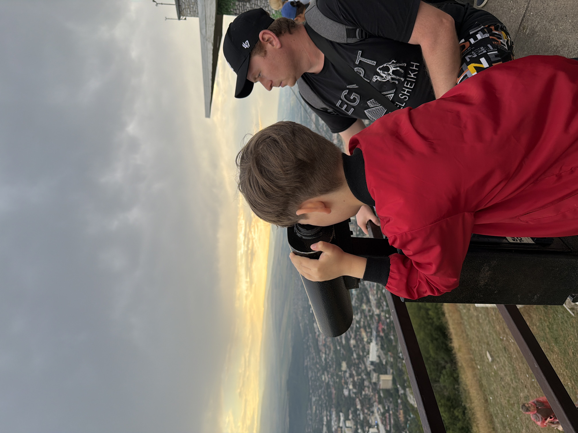 An overcast, late afternoon sky looms over a city panorama viewed from a high vantage point. In the foreground, a young boy, wearing a bright red jacket, is engrossed in looking through a coin-operated telescope. To his right, a man in a black baseball cap and a black t-shirt with graphics of camels and Egyptian pyramids is seated and looking in the same direction as the boy. The man wears a backpack. The telescope is mounted on a dark, heavy base, and a metal railing runs along the edge of the viewing platform. Below the platform, a grassy slope descends towards the city, with scattered buildings visible in the distance, extending towards a hazy horizon. Another person wearing a red poncho can be seen walking on the slope. The overall impression is of tourists observing a scenic view of Goryachevodsky, Russia.