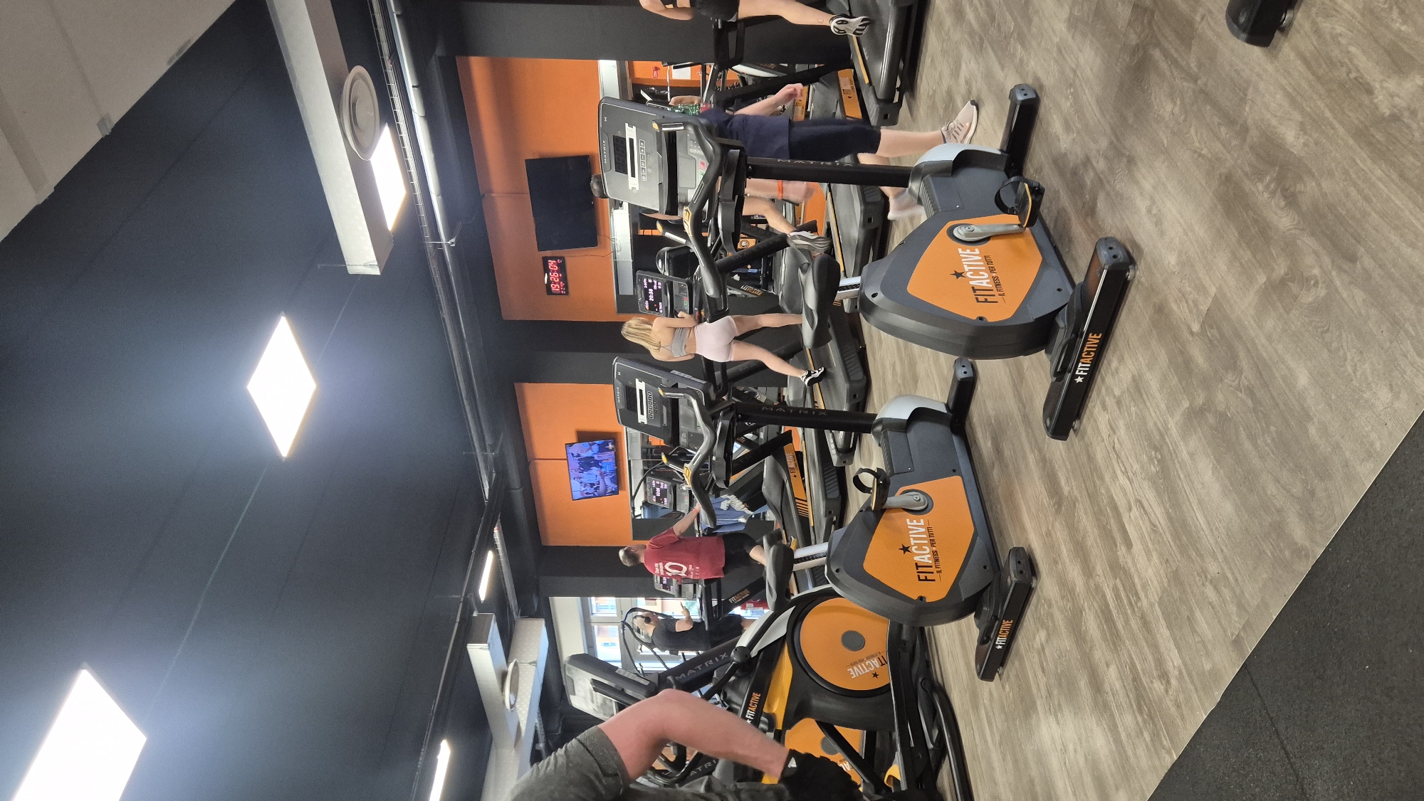 This is a photograph of the interior of a gym. The scene is filled with various exercise machines, including treadmills and elliptical trainers. Several people are visible, engaged in their workouts. One woman in a grey sports bra and pink shorts is on a treadmill. Another person in a red t-shirt is on an elliptical machine. The gym features orange accents on the equipment and walls, contrasting with the dark ceiling and grey floor. The lighting appears to be artificial, with bright square lights on the ceiling. Text visible on the equipment includes "FITACTIVE" and "MATRIX". A digital clock on the wall displays "19:26:04", suggesting the time of day is in the evening. The overall atmosphere seems to be one of active exercise.