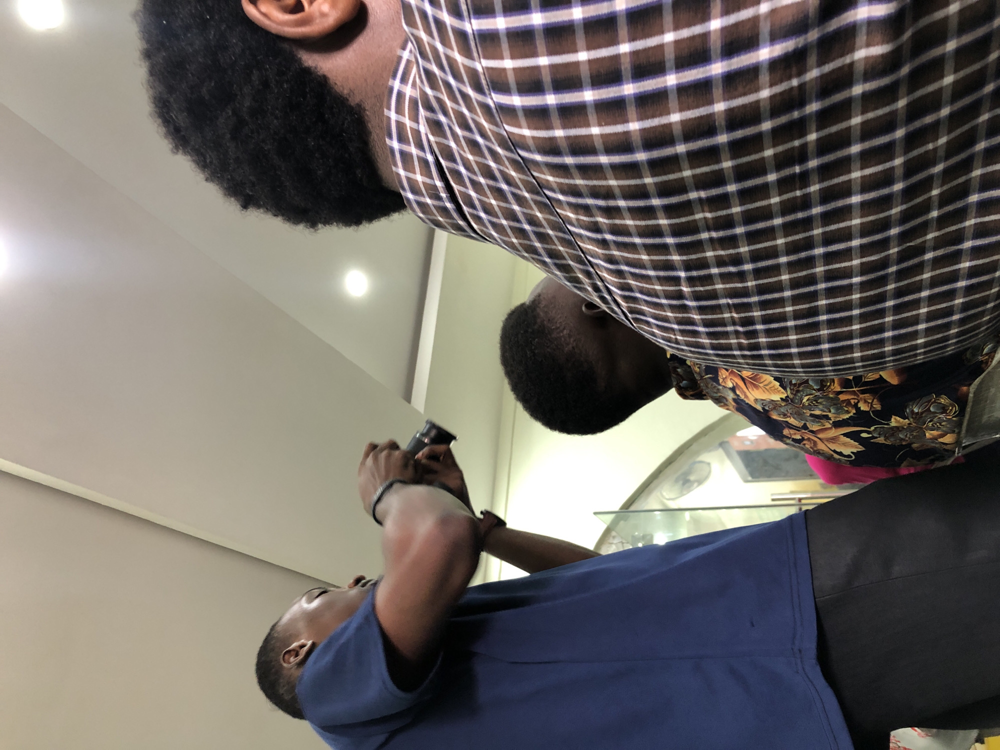 In Ikeja, Nigeria, a man in a blue t-shirt holds a camera and appears to be taking a picture or video. He is positioned to the left of the frame, facing away from the viewer. To his right, another person, partially visible, is wearing a brown and white checkered shirt, their back to the camera. Between these two individuals, a third person's head is visible, facing down. The background suggests an indoor setting with a white ceiling and light fixtures. The overall impression is of a casual gathering or event where photography is taking place.