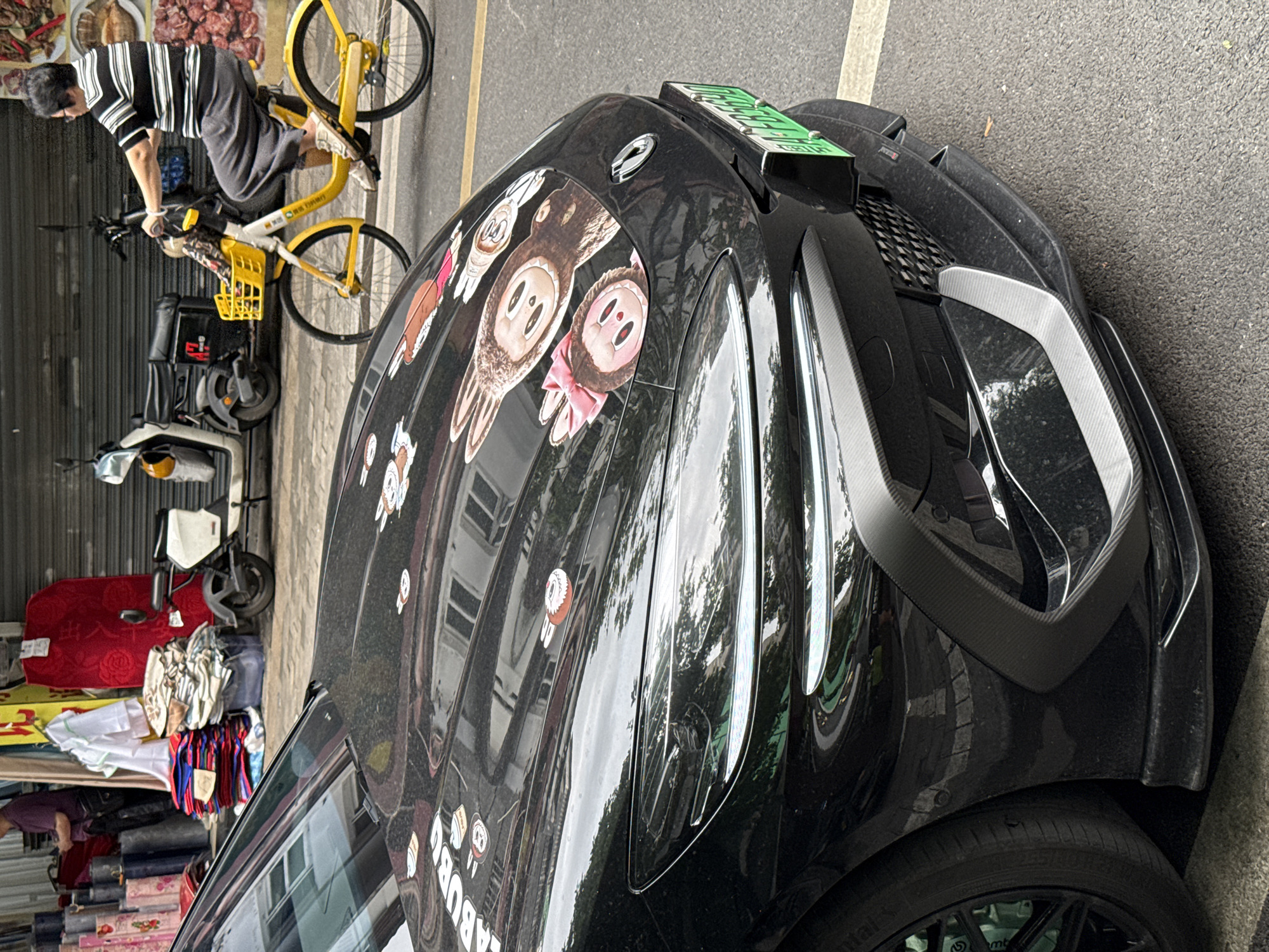 A black car covered in cartoon-like animal stickers parks on a Liwan District, China street. A man wearing a striped shirt rides a yellow bike next to the car, with another bike and a scooter also nearby. A store next to the bike and scooter sells various fabrics and goods. The environment suggests a bustling commercial area within the urban context of Liwan District.