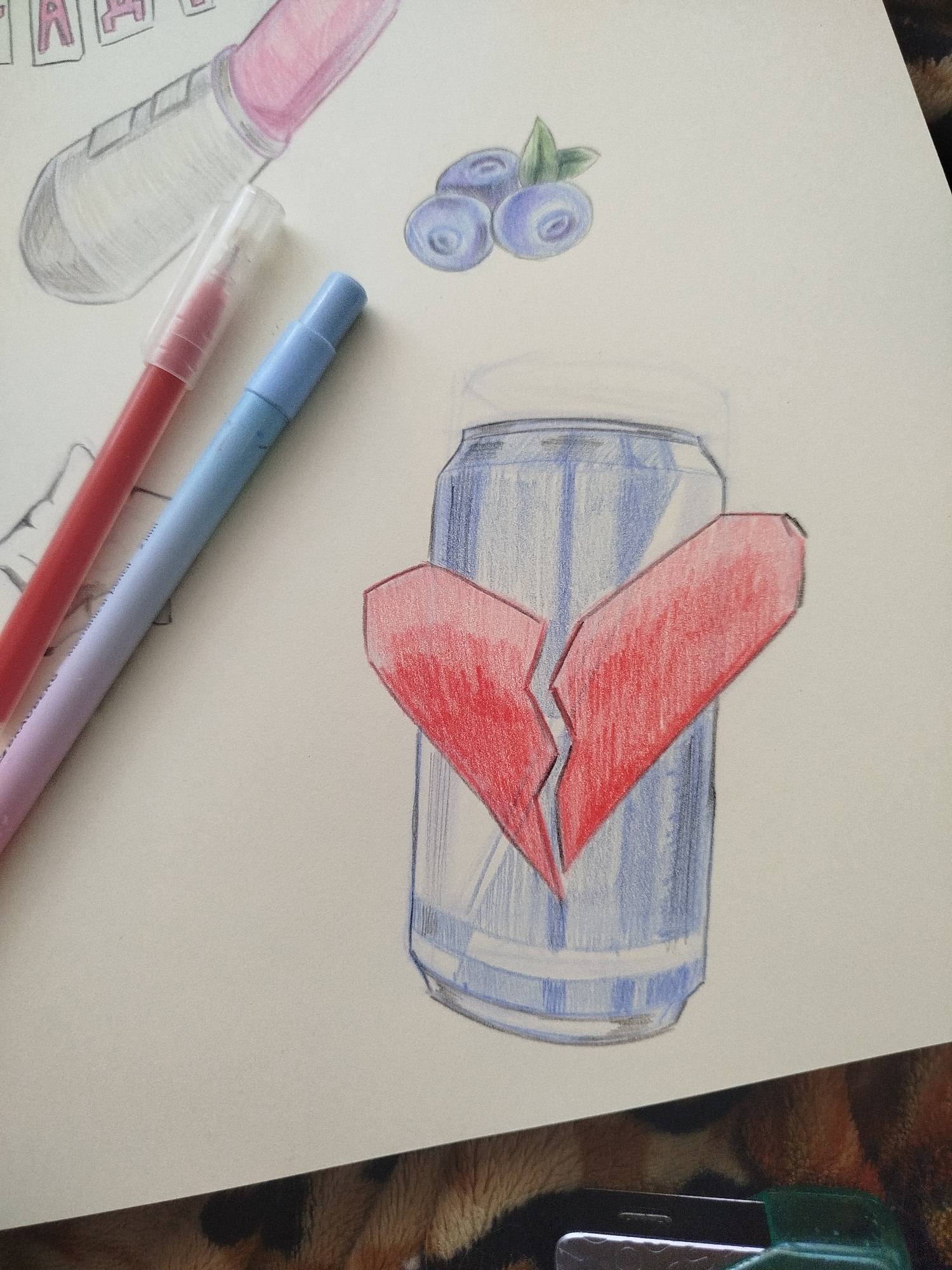 The image is a drawing on paper, depicting a broken heart over a blue can. Above the can are three blueberries with green leaves. To the upper left are a drawing of a tube of lip gloss and some letters. Two pens, one red and one blue, are laying to the left of the can. A portion of a stapler and animal-print cloth are visible in the lower right.