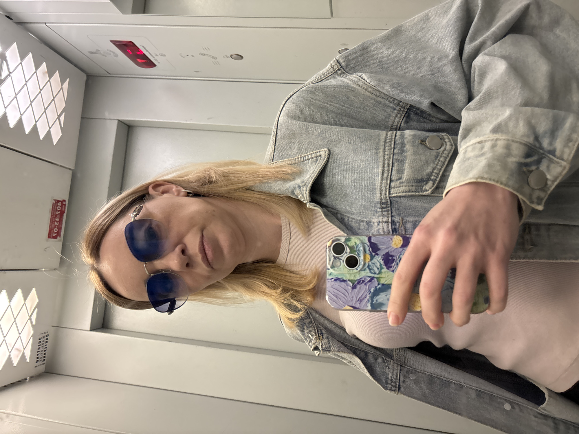 This image is a selfie of a person, likely female, taken in what appears to be an elevator or a similar enclosed, box-like space. The image is rotated 90 degrees clockwise, showing the person on their side.
The subject has shoulder-length blonde hair and is wearing blue-tinted sunglasses, which obscure part of her face. She is dressed in a light blue denim jacket over a light-colored, possibly pink or white, ribbed top. Small silver earrings are visible, one appearing to be a safety pin and another a small charm. She holds a smartphone with a distinctive floral-patterned case (featuring shades of purple, green, yellow, and white abstract flowers) in her right hand, angled towards herself, indicating a selfie. Her expression is neutral.
The setting consists of light-colored, possibly metallic walls and ceiling. On the left side, there's a decorative, backlit panel with a grid of diamond-shaped cutouts. Above, a red illuminated digital display is visible. The ceiling also features small, recessed lights.
Notable details include the bright, artificial lighting within the confined space. On the red digital display, faint, blurry text resembling "LIF" or "LIFT" and possibly a number like "10" can be seen. Below the diamond-patterned panel, a red sticker with Cyrillic text is partially visible, showing fragments like "РЭ-ТО-ОТ" or "НЕ ТО," and words such as "СВЯЗЬ" (connection) and "ЭЛЕКТРИК" (electrician) or related terms.