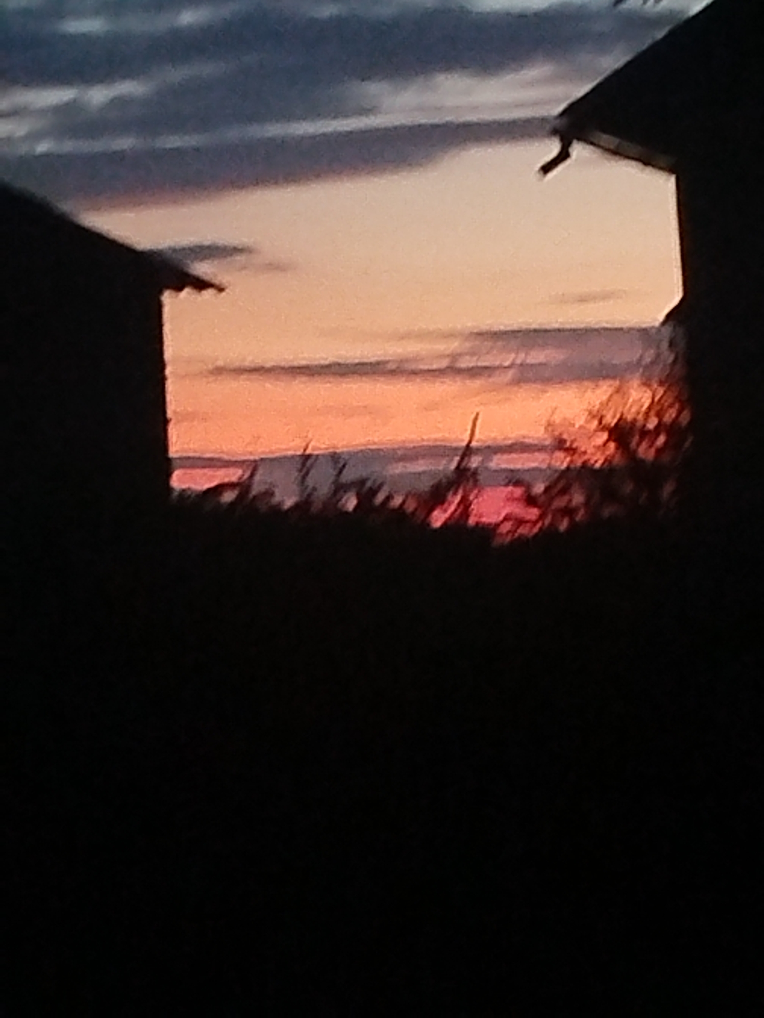 The image captures a twilight scene from Zastavna, Ukraine. Two silhouetted structures, likely buildings, frame the view. One is on the left, partially obscuring the sky and the other building on the right. The sky displays a gradient of colors, transitioning from dark gray clouds at the top to vibrant orange and pink hues near the horizon, indicating a sunset or sunrise. In the foreground, a dark silhouette of vegetation or trees is visible, with some indistinct shapes that might be distant structures or elements of the landscape. The overall impression is one of a calm, natural setting during the transition between day and night, framed by human-made structures.