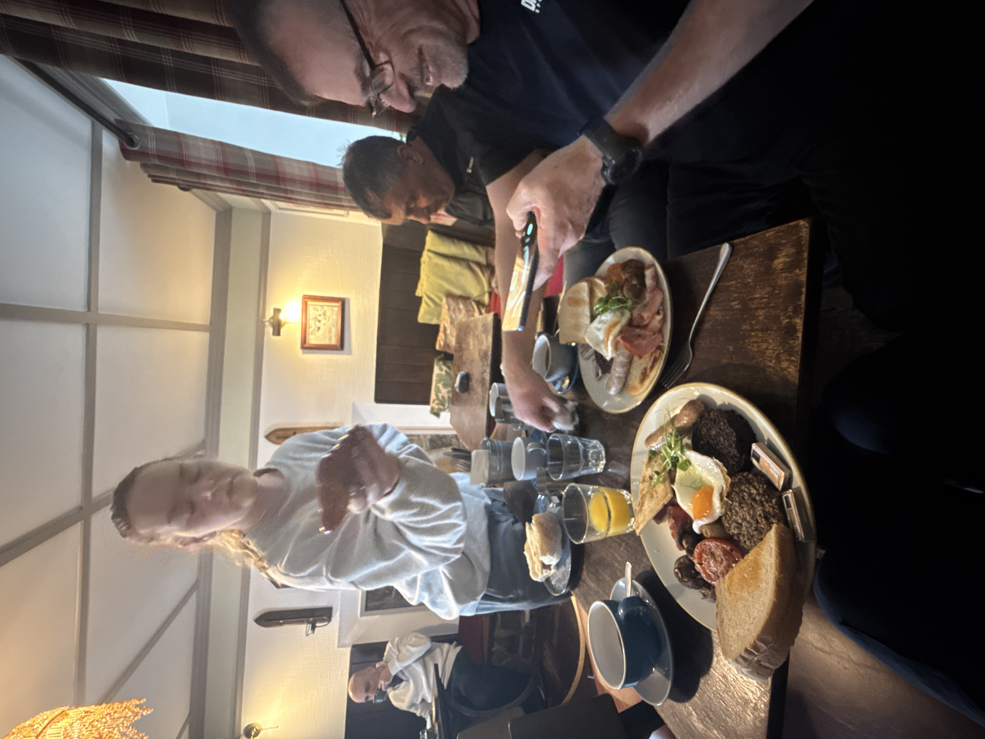 A group of people are sitting around a wooden table in what appears to be the inside of a pub or restaurant in Unknown, Unknown. The table is set with plates of food, including a full English breakfast with items like sausages, black pudding, eggs, tomatoes, and toast. There are also mugs and glasses on the table, some containing orange juice. The room is illuminated by a wall sconce and natural light coming from a window. The window is adorned with a curtain. Some people are interacting with the food, and one person seems to be taking a picture with their mobile phone.