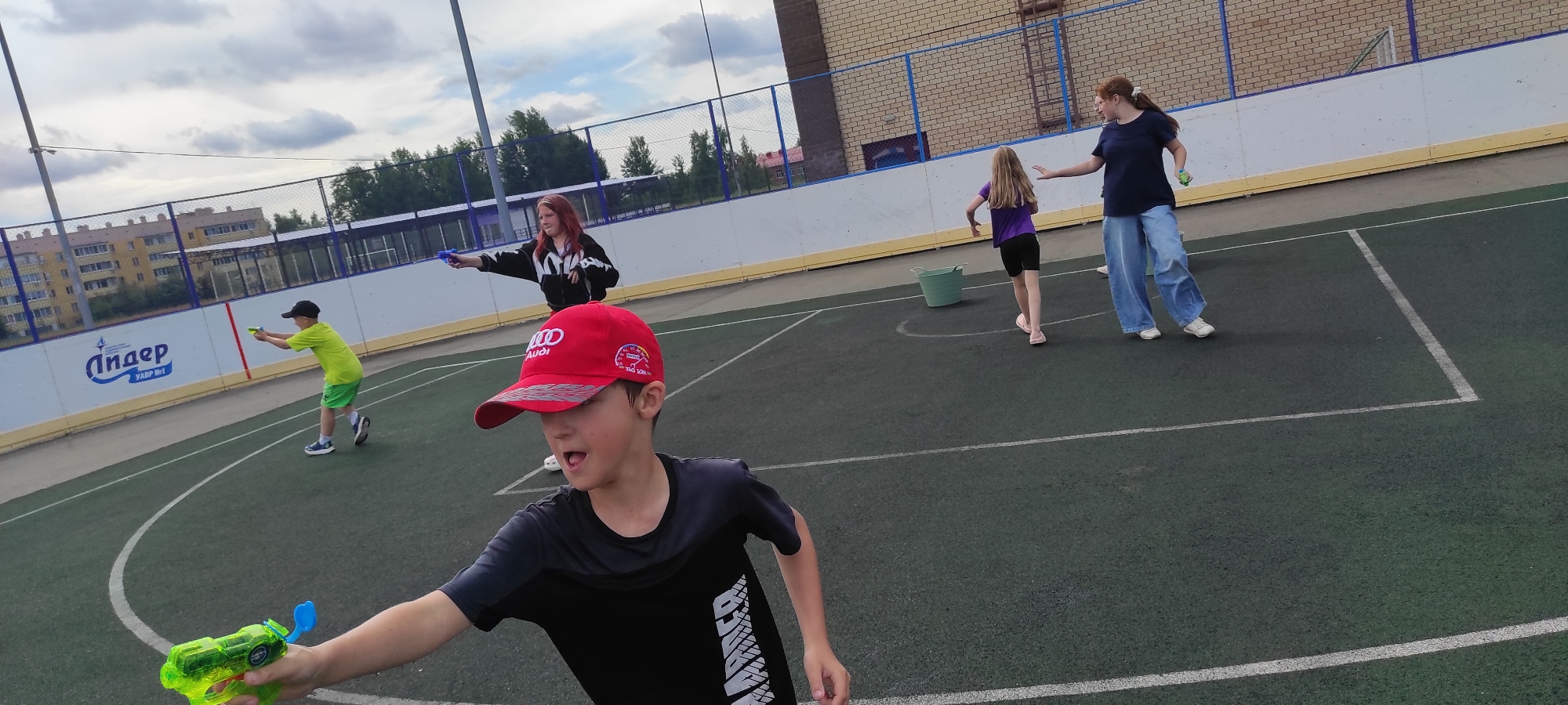 In Noviy, Russia, on a cloudy day, a group of four people, including two children and two young women, are engaged in a water gun fight on a dark green artificial turf sports field. The field has white boundary lines and is enclosed by a blue fence. In the foreground, a boy wearing a black t-shirt and a red baseball cap with the Audi logo is holding a neon green water gun and appears to be mid-action, with his mouth open. Behind him and to the left, a younger boy in a bright green t-shirt and shorts is also aiming a yellow water gun. In the middle ground, a young woman with red hair, wearing a black and white hooded jacket, aims a blue water gun. Further back and to the right, another young woman in a dark blue t-shirt and baggy blue jeans is looking towards the children and holding a small green object, possibly another water gun or a projectile. A light green bucket is situated near the center of the field. In the background, a multi-story apartment building and trees are visible beyond the fence, suggesting an urban or suburban setting.