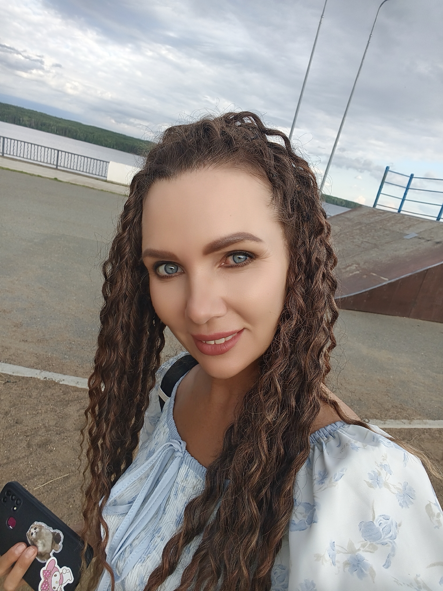 A selfie of a woman with long, curly brown hair and blue eyes is taken outdoors with a cloudy sky. She is smiling and holding a black smartphone adorned with stickers. She is wearing a light blue off-the-shoulder top with a floral pattern. In the background, a body of water, a railing, a road, and a ramp structure are visible. The location is the embankment on the Kama River in the city of Chaikovsky, Russia.