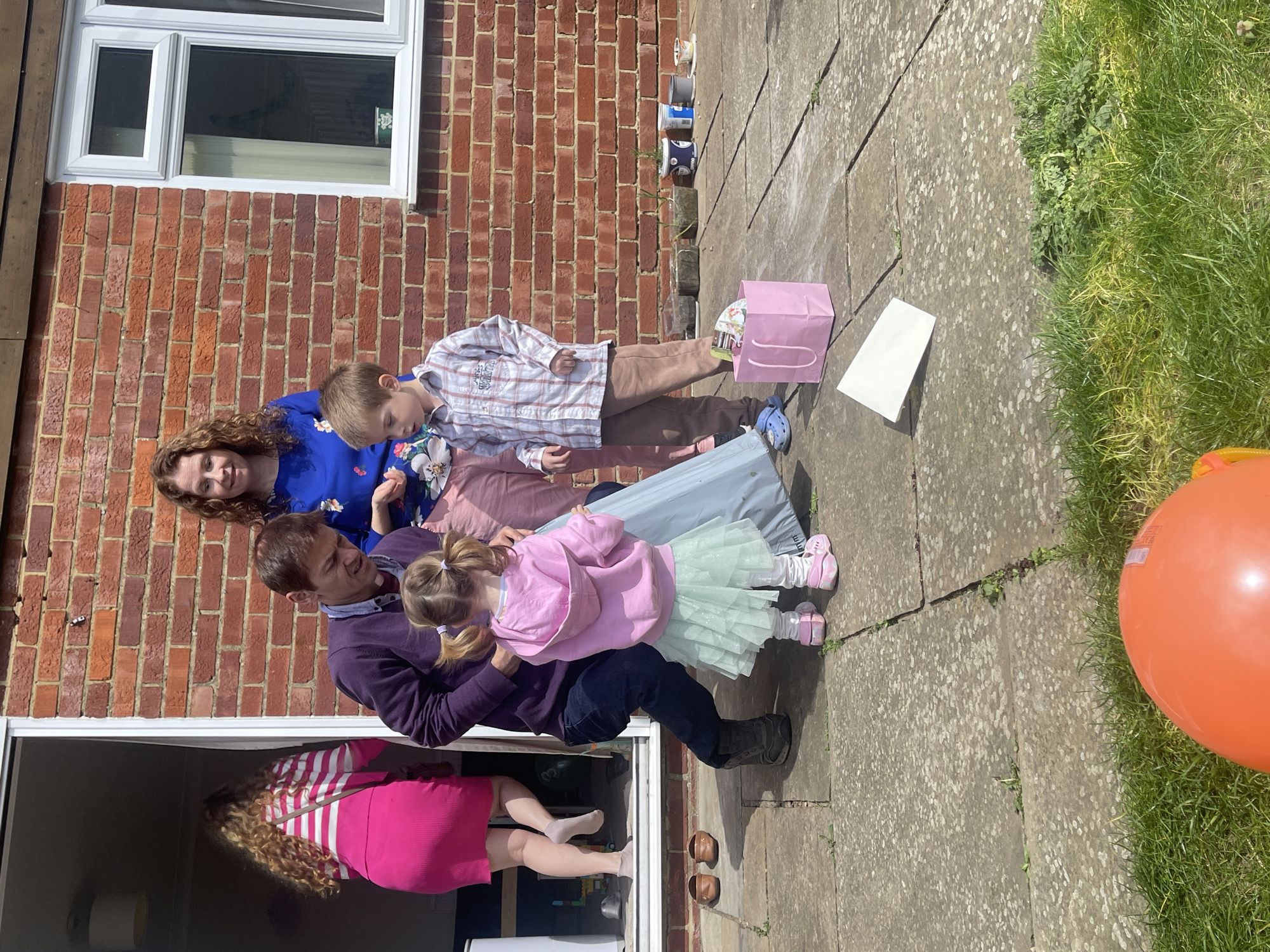 The image captures a family gathering outdoors, likely a birthday celebration, in what appears to be a backyard patio setting. A group of five people are visible: a woman with curly hair wearing a blue floral-patterned top, a boy in a plaid shirt and brown pants, a child in a pink hooded sweatshirt and a green tutu, a man in a purple sweater and jeans, and a woman in a pink and white striped dress and blonde hair. The man is holding the young girl in the tutu. The location shows a brick exterior wall with a window and a patio paved with stone tiles. A pink gift bag and a white envelope are placed on the patio floor. A row of plant pots lines the wall. A bright orange balloon is visible in the grass. The location is likely in Maidstone, United Kingdom.