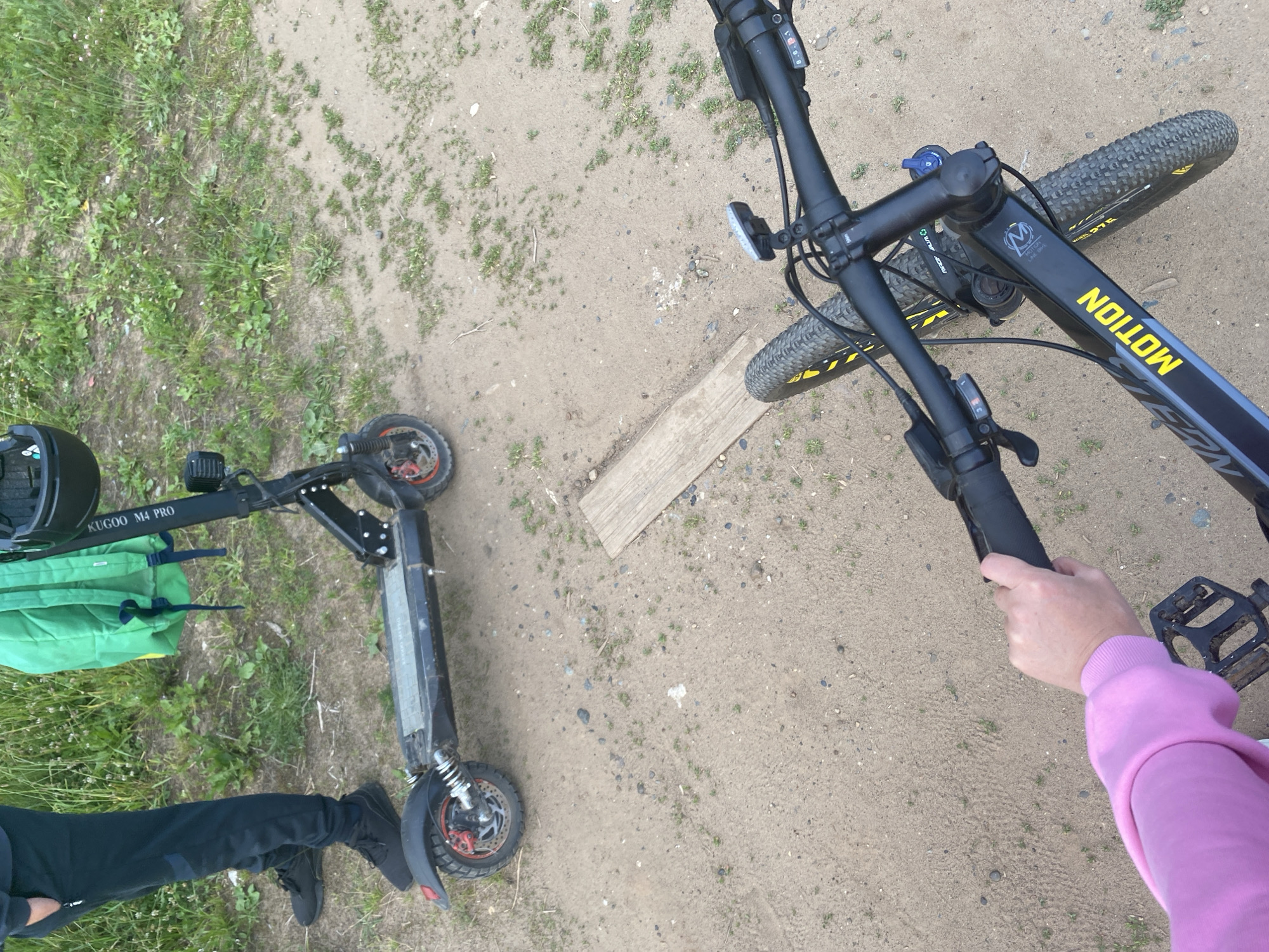 The image shows a top-down view of two electric scooters and a bicycle on a dirt and grass surface. One scooter, identified by the text "KUGOO M4 PRO" on its frame, is black with a green bag draped over its handlebars. A person's legs, clad in black pants and shoes, are visible next to this scooter. The second scooter is also black. A bicycle with a black frame and yellow accents, bearing the brand name "MOTION" in yellow, is positioned to the right. The handlebars and front wheel of the bicycle are clearly visible, with a person's hand wearing a pink sweatshirt holding onto the handlebars. The scene appears to be outdoors, possibly in a park or recreational area, with dry dirt and patches of green grass. The lighting suggests it is daytime. There are no strong indicators of weather conditions, but the ground appears dry. The overall impression is one of a casual outdoor setting, perhaps before or after an activity involving these vehicles. The image is taken in Chaikovsky, Russia.