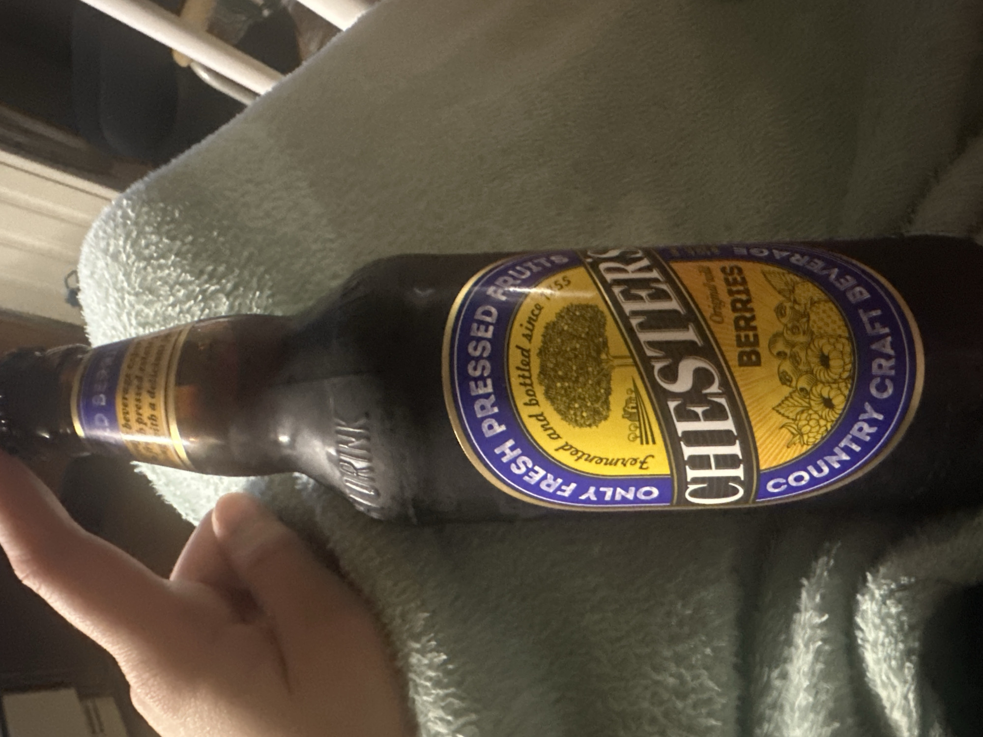 The image shows a bottle of "Chester's Country Craft Beverage" resting on a soft, light-colored surface, possibly a blanket or towel. The bottle is dark brown and features a label with a blue and yellow design that reads "Only Fresh Fermented and bottled since 1955" and "Pressed Fruits". It also has the word "Chesters" prominently displayed in a large font and the word "Berries" written next to it. A person's hand is partially visible near the bottle's neck. The background is blurry and dimly lit, with some unidentified objects. The scene is not in Moscow, Russia.