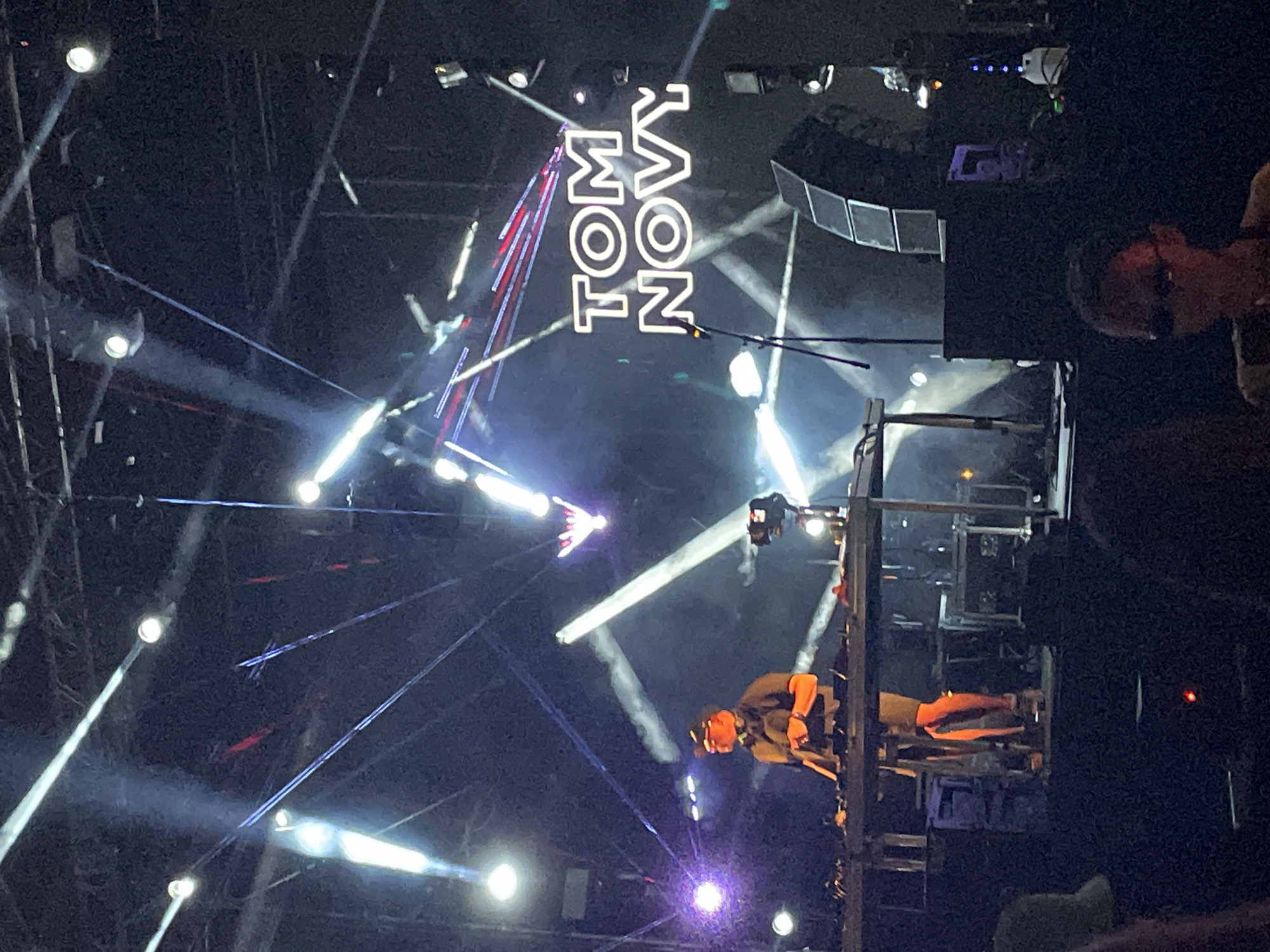 The image depicts a concert stage with lighting and a performer in Bar, Montenegro. The band "TOM NOVY" is emblazoned in white lettering above the stage. The performer is elevated on a structure, illuminated by beams of light. Other lights, rigging, and equipment are visible, creating a dynamic stage environment.