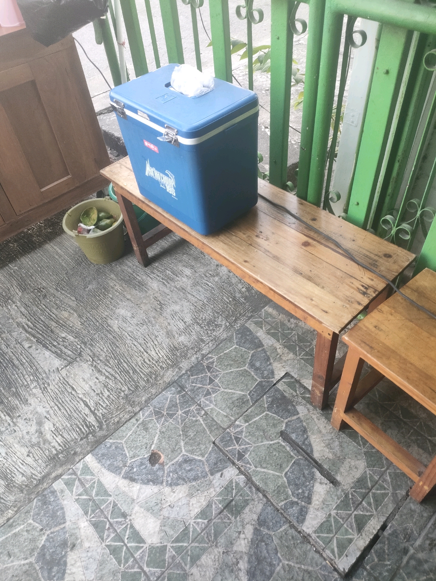 A high-angle outdoor shot shows a blue cooler on a wooden bench in front of a green metal fence. To the left, a portion of a wooden cabinet is visible. In front of the cooler, a yellow bucket contains several green fruits, possibly avocados. The ground is a mix of textured concrete and patterned tiles. The scene appears to be in a residential or public area in Surabaya, Indonesia.