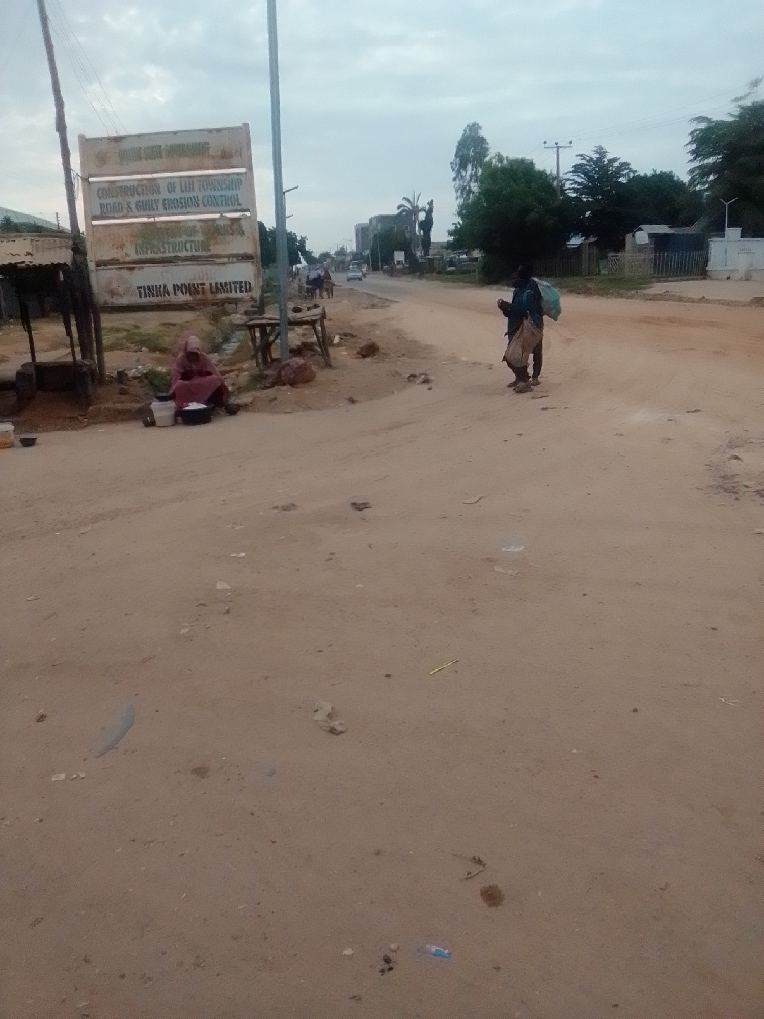 In the city of Gombe, Nigeria, a dirt road is depicted under a cloudy sky. A large, weathered signpost stands on the left side of the road, displaying text related to road and gully erosion control projects by Tinka Point Limited. Next to the signpost, a person is seated on the ground, surrounded by pots and containers. Further down the road, another individual walks away from the camera, carrying bags over their shoulder. Buildings, trees, and utility poles line the background. The ground is dry and sandy, with scattered debris.