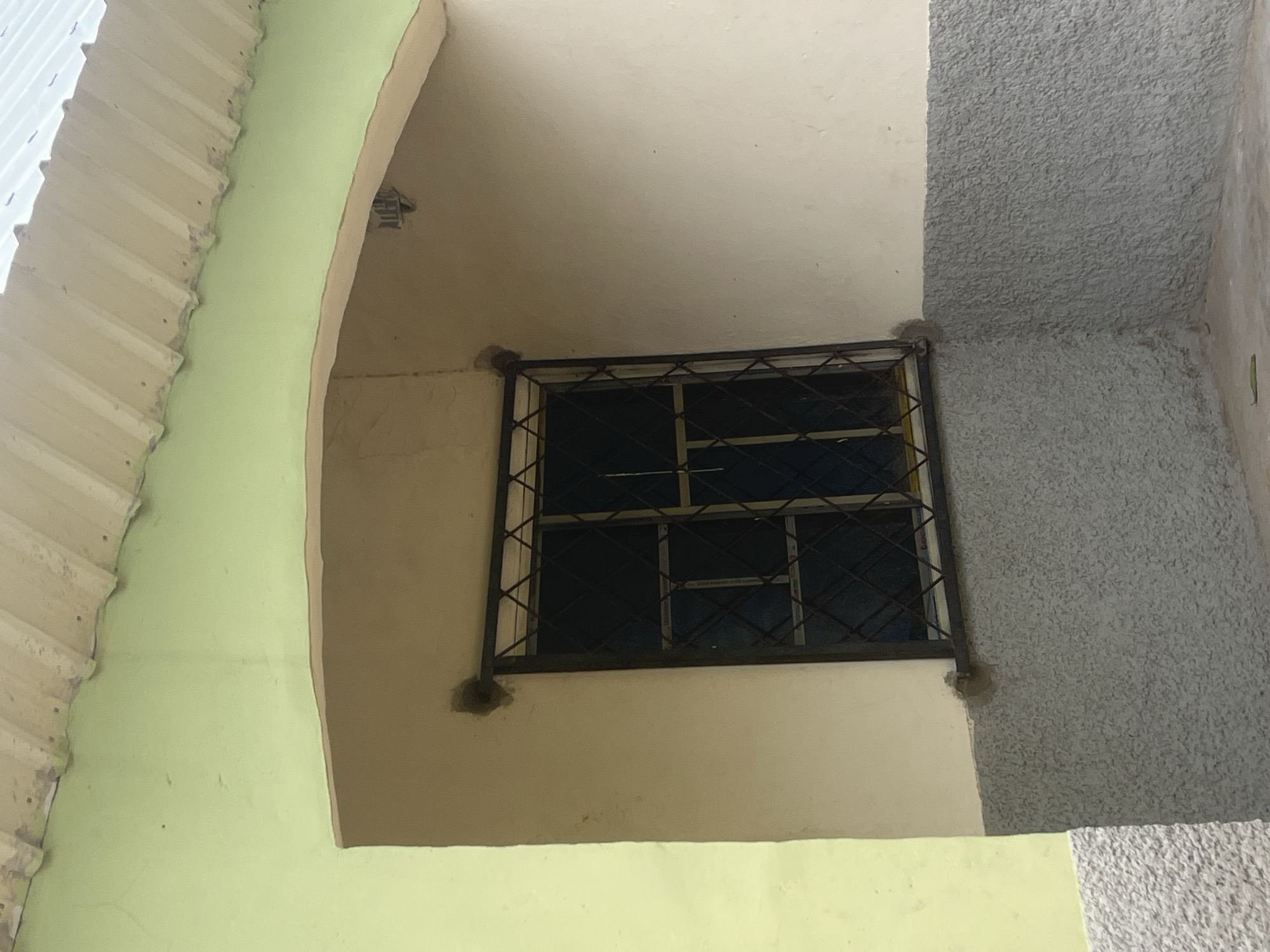 The image shows a close-up view of the exterior of a building with a light green upper wall and a grey, textured lower wall. A window with a dark metal grate is set into the wall, recessed within an arched alcove. The roofline, partially visible at the top of the image, appears to be made of corrugated metal. The scene is likely in Jalingo, Nigeria, given the context provided. There are no people or discernible actions in the image; it focuses on the architectural details of the building.