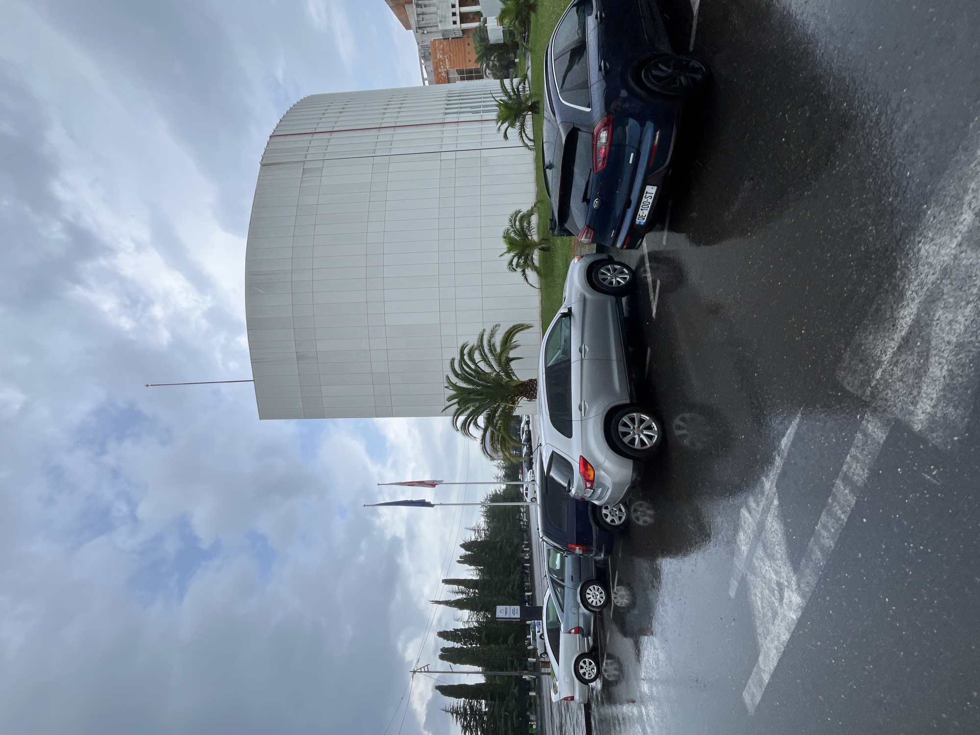 The image depicts a parking lot in front of a modern, curved white building, likely in the city of Poti, Georgia. The ground is wet, indicating recent rainfall, and is marked with white parking lines. Several cars are parked, including a silver SUV in the foreground, a dark blue SUV behind it with a license plate "DE-100-ST," a dark blue minivan, a silver sedan, and a light silver sedan at the far left. In the background, there are palm trees, cypress trees, and flags flying from poles in front of the building. The sky is cloudy, with patches of blue visible. The overall scene suggests an overcast day in a coastal or southern European-style city.