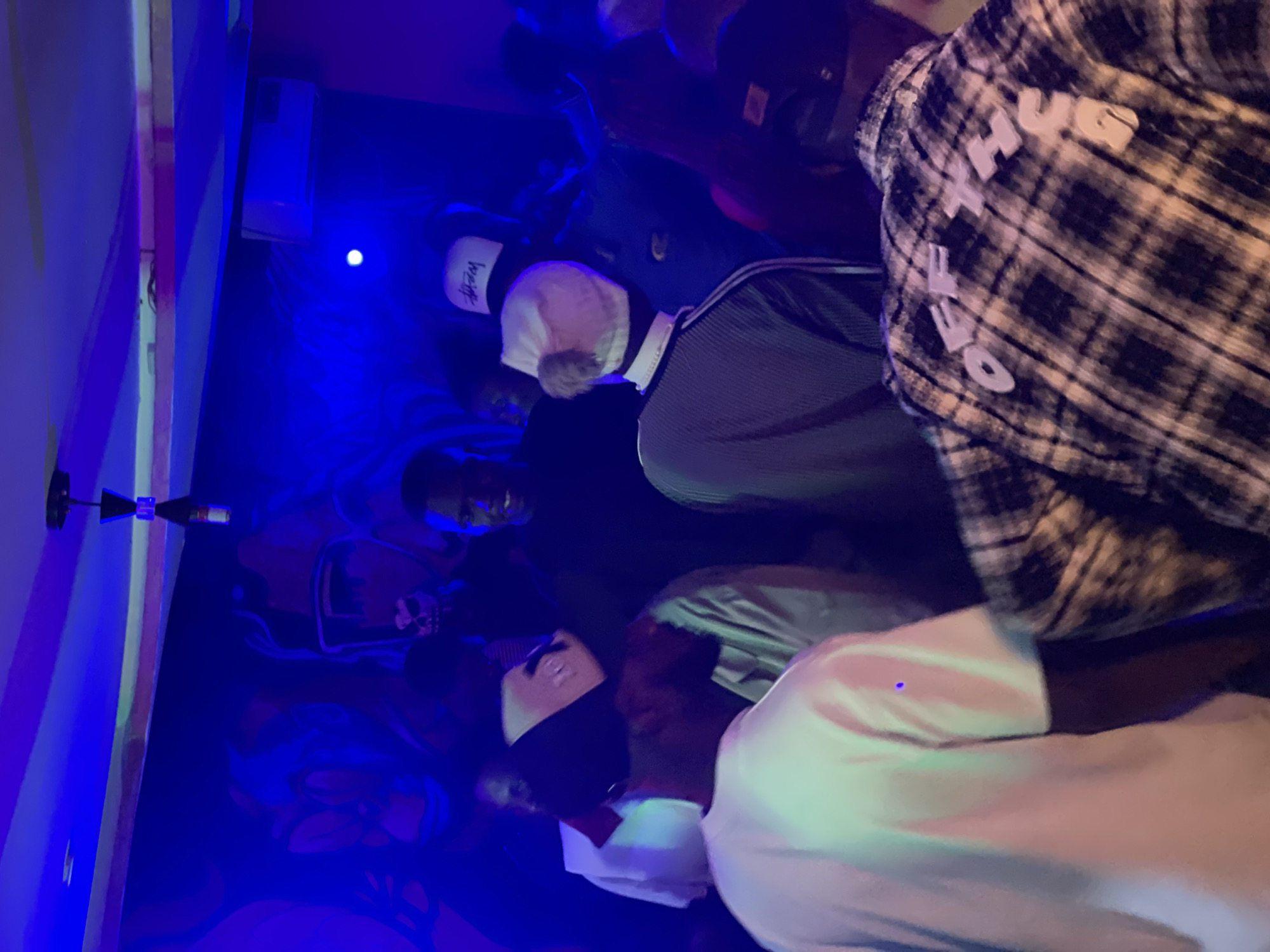 The image depicts a group of people inside a dimly lit indoor venue, likely a club or lounge, in Durumi, Nigeria. The space is illuminated by vibrant blue and purple lights, creating a club-like atmosphere. A mural, featuring stylized faces and possibly skulls, adorns the wall in the background, further enhancing the ambiance.
In the foreground, a person wearing a white t-shirt is visible from the back, their arm outstretched. To their right, another individual is seen in profile, wearing a black and white plaid shirt with the text "OFF THE US" emblazoned on the back. This individual appears to be interacting with someone just out of frame.
Behind these figures, several other people are present, some wearing hats. One person in the center is looking towards the camera, smiling. The overall impression is of a social gathering in a nightlife setting.