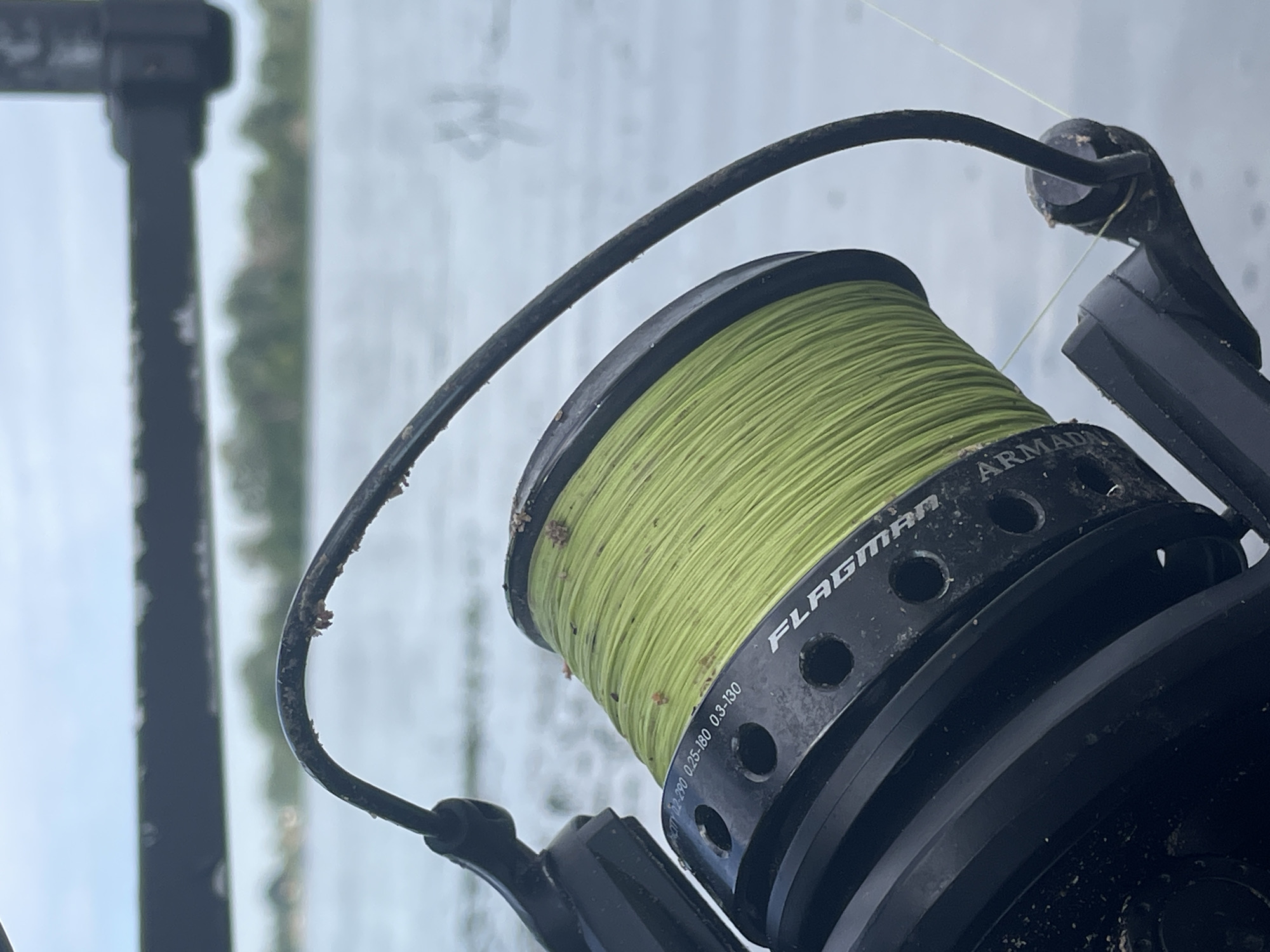 The image shows a close-up of a fishing reel with bright green fishing line wound around it. The reel is black and has some lettering that appears to say "FLAGMAN ARMADA" and "CAPACITY 0.2-290 0.25-180 0.3-130". The reel is partially covered in dirt or sand. A black metal rod extends horizontally across the top of the image. In the background, there is a body of water, likely a river or lake, with trees and buildings visible on the far bank under a cloudy sky. The overall scene suggests an outdoor fishing setting. The provided context places this image in Kyiv, Ukraine, implying the water body is likely the Dnieper River.