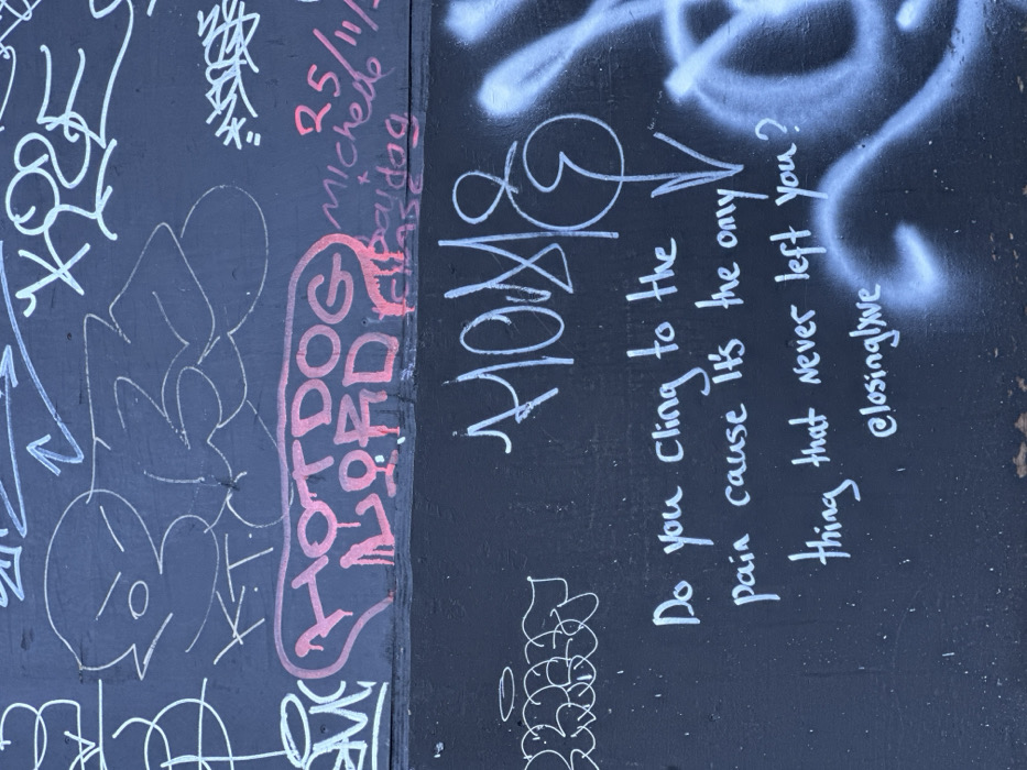 A dark-colored wall or hoarding, divided by a horizontal seam, is covered with various graffiti tags and artwork. The upper section features numerous white outlines, including abstract shapes, arrows, and a stylized character with angry eyes and a circular mouth, positioned next to the number '3'. Below these, the words "HOTDOG LORD" are painted in red bubble letters with visible drips. To the right, faint red script includes a date ("25/11/..."), "Michelle", "paidog", and partially visible "ease". The lower section of the wall displays a large, stylized white tag, possibly reading "HOME" or "MOM&", which incorporates a downward-pointing arrow. Below this, a white handwritten message states: "Do you cling to the pain cause it's the only thing that never left you?", followed by "@closinglxve". To the right, a cloud of light blue and white spray paint partially obscures other faint markings. This scene is located in the city of London, United Kingdom.
