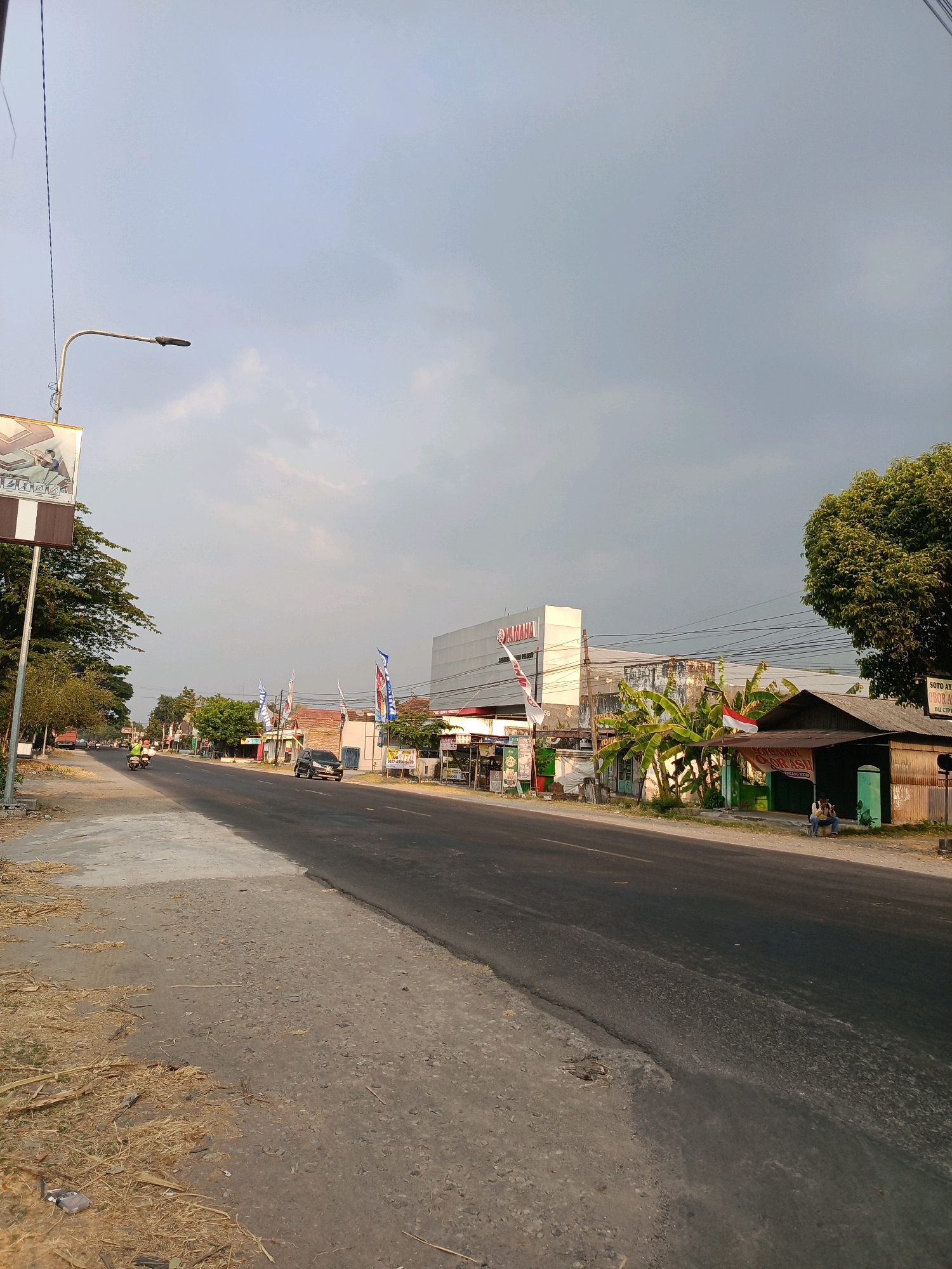 The image depicts a street scene in Kincang Wetan, Indonesia, under a cloudy sky. The road, a prominent feature, is paved with asphalt and runs diagonally from the lower left to the upper right of the frame. To the left of the road, a dirt and gravel shoulder is visible, scattered with dry vegetation.
Along the right side of the road, several buildings and structures are present. A large white building with the "YAMAHA" logo prominently displayed stands out. Adjacent to it is a smaller, older building with a corrugated metal roof and a green facade. Banana trees are planted in front of these buildings, and a red and white Indonesian flag waves from a pole. Further down the road on the right, a small food stall with a wooden structure and a tin roof is situated, with two individuals sitting on a bench outside.
Several vehicles are on the road, including a black car and a motorcycle with two riders. Vertical banners or flags with various graphics are placed along the roadside, some appearing to be advertisements. A street lamp is visible on the left side of the road, and telephone wires stretch across the sky. Trees and vegetation line both sides of the street, creating a somewhat rural or suburban atmosphere within the town. The overall lighting suggests it is either early morning or late afternoon.