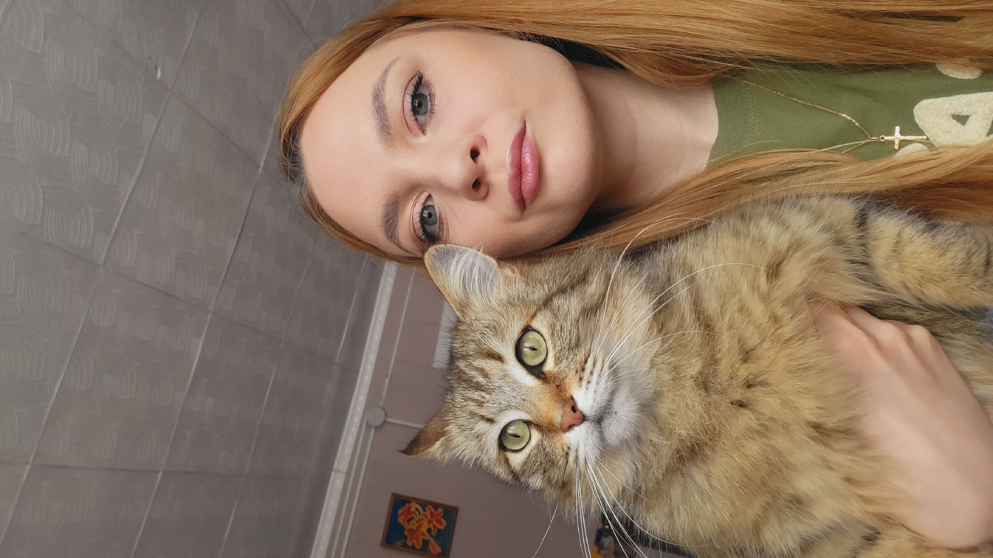 A young woman with light red hair and pale skin holds a brown tabby cat. The woman wears a green shirt and a gold necklace with a cross pendant. The cat has green eyes and a long white whiskers. The scene appears to be indoors, possibly in a home, as evidenced by the grey tiled wall and a framed picture hanging on the wall in the background. The exact location is Зипуново, Russia.