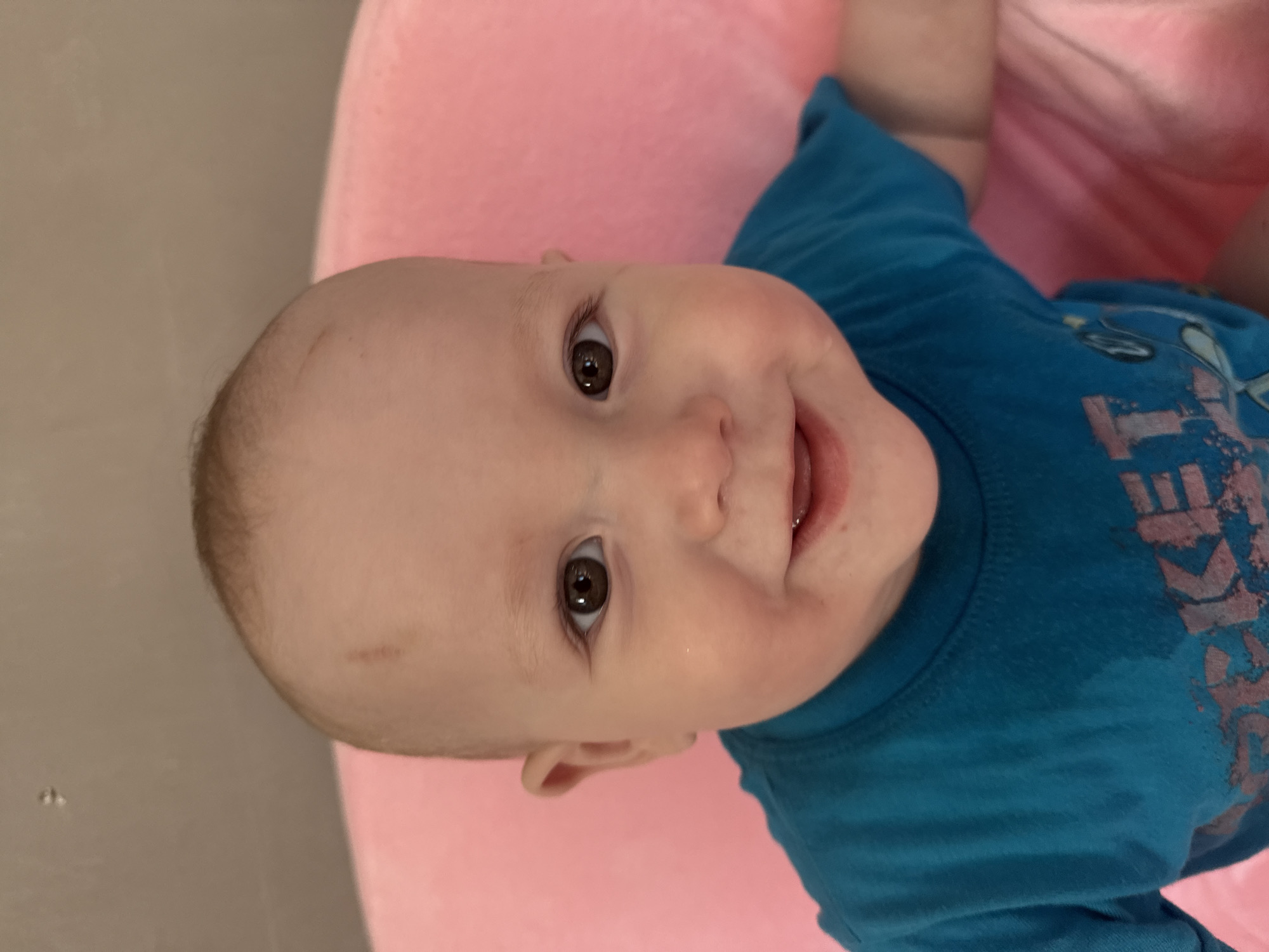 A close-up, head-and-shoulders portrait depicts an infant, estimated between six and eighteen months of age, with fair skin, dark brown eyes, and sparse light-brown hair. The infant is looking directly at the viewer with a broad smile, mouth slightly open, revealing the lower gum line. A faint, reddish-brown linear mark is visible on the upper left forehead, above the eyebrow. The infant is wearing a teal blue short-sleeved t-shirt, on which a faded graphic design is partially visible, including the letters "ROCKET". The infant appears to be seated against or surrounded by a soft, textured pink object, possibly a cushion or chair. A plain, light beige or taupe wall serves as the background in the upper portion of the image. The scene is located indoors, in the city of Кипчак, Russia.